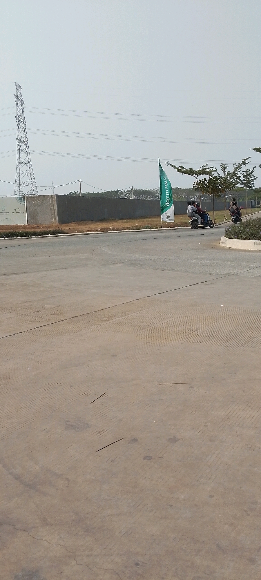 In Satriamekar, Indonesia, this image captures a scene on a clear, bright day. The foreground is dominated by a paved surface, likely a road or pathway, with faint lines and patterns visible. In the midground, a curved road leads towards a development area. To the left, a large concrete wall forms a boundary, with signage indicating "SUTRACITRA LAND" and a logo. A tall electrical transmission tower is visible in the far left background, along with power lines stretching across the sky. To the right of the wall, a green flag with white lettering that reads "darmawa" stands prominently. Two individuals are seen riding on a scooter, with one passenger perched behind the driver. Further to the right, another individual is on a separate scooter, also moving along the road. Several trees with sparse foliage are scattered throughout the midground and background, suggesting a semi-developed or developing area. The overall environment appears to be an outdoor space with elements of infrastructure and construction.