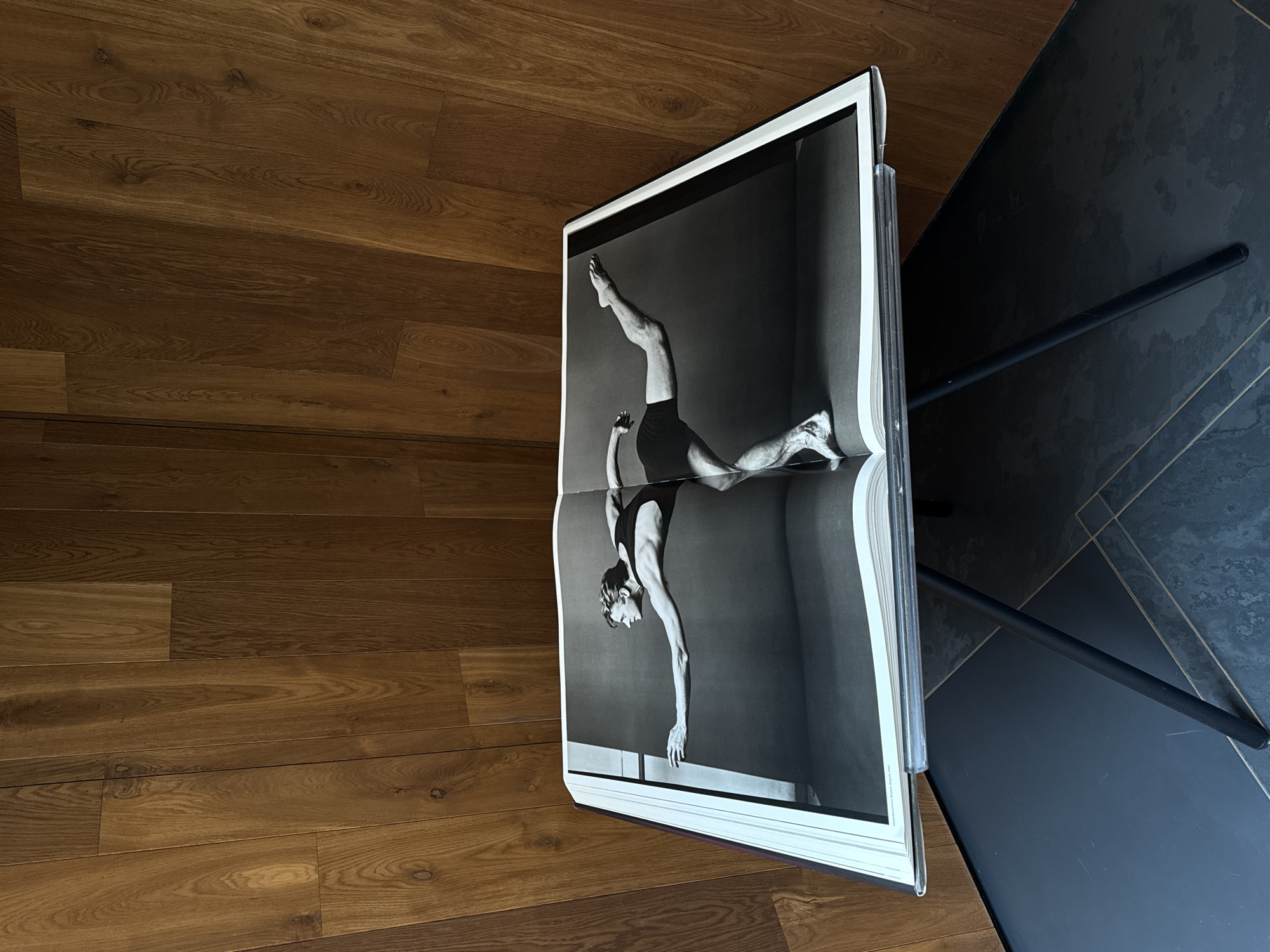 An open, large format book rests on a minimalist black stand with thin, angled metal legs. The book is splayed open to a two-page spread featuring a black and white photograph of a muscular male dancer. The dancer is depicted in a dynamic, balanced pose, barefoot and wearing a dark, form-fitting garment, with his left leg extended backward and upward, and his left arm reaching forward. The background of the photograph is a plain, dark studio setting. The stand is positioned on a dark tiled floor that displays a subtle geometric pattern with light lines. Behind the stand and book is a vertical wooden wall composed of individual planks, exhibiting various natural brown tones and grain textures. The scene is indoors. No specific location within Mexico City is discernible from the visual information provided.