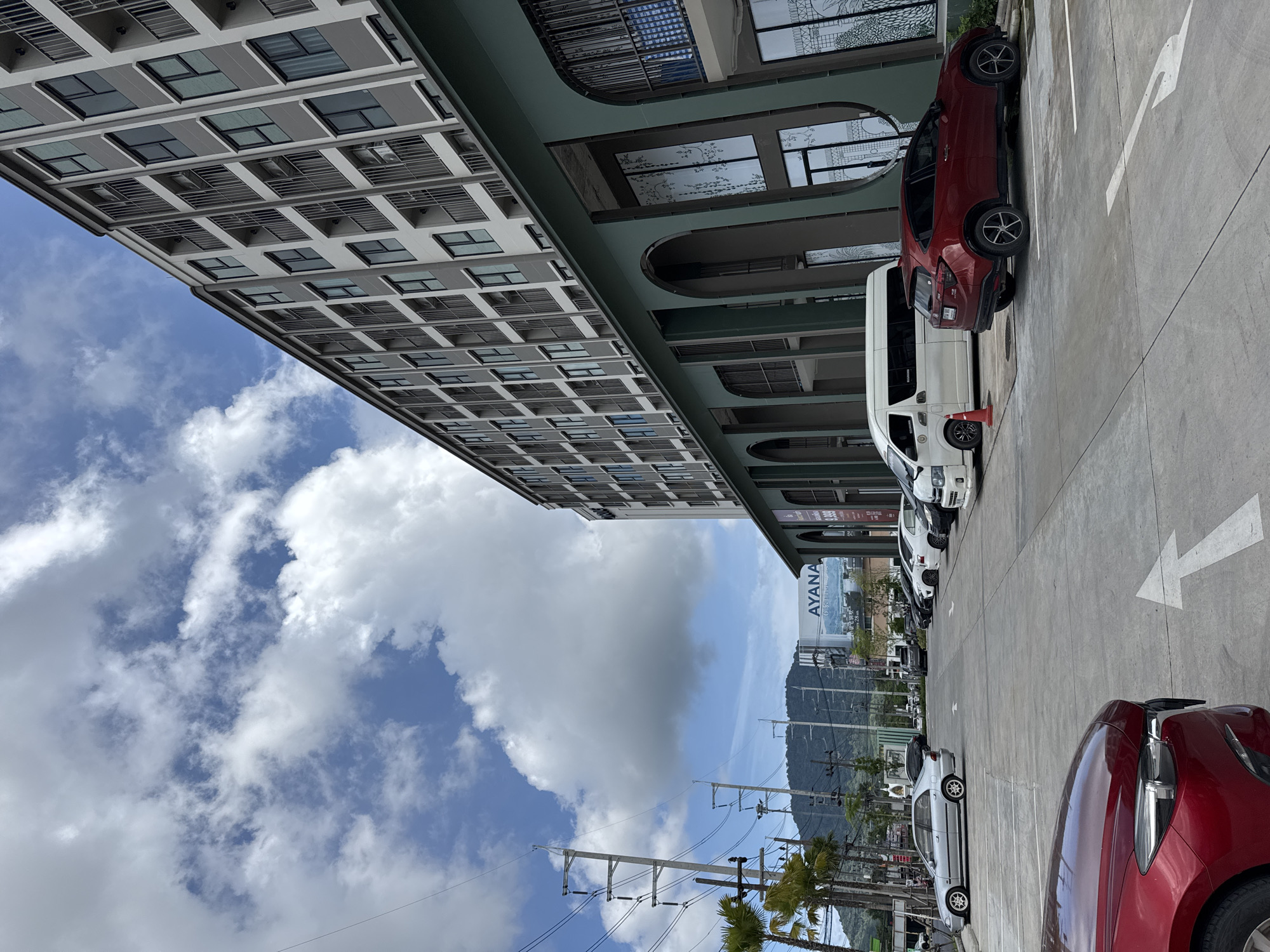 This is a medium-angle, eye-level, full exterior shot of a modern apartment building's ground floor with an adjacent parking lot. The building features a repeating pattern of dark green arched openings and large windows, with balconies and windows on the upper floors. In the foreground, there is a red car parked in a lot with white directional arrows painted on the asphalt. A white van and a red SUV are also visible in the parking lot. Beyond the parking lot, there is a street with power lines and palm trees. In the background, a large billboard advertising "AYANA" is visible in front of a mountain. The sky is blue with scattered white clouds. The scene is located in Kathu, Thailand.