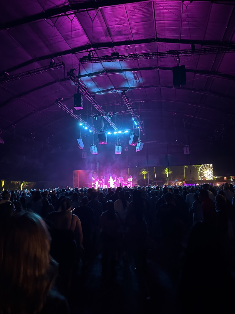 The image depicts a concert at night, likely in a large tent or covered venue in Indio, United States. A crowd of people faces a brightly lit stage, where a band or performers are visible. Lighting rigs hang above the stage and the crowd. In the distance, palm trees and a Ferris wheel are visible. The scene is dimly lit with purple and blue stage lights.