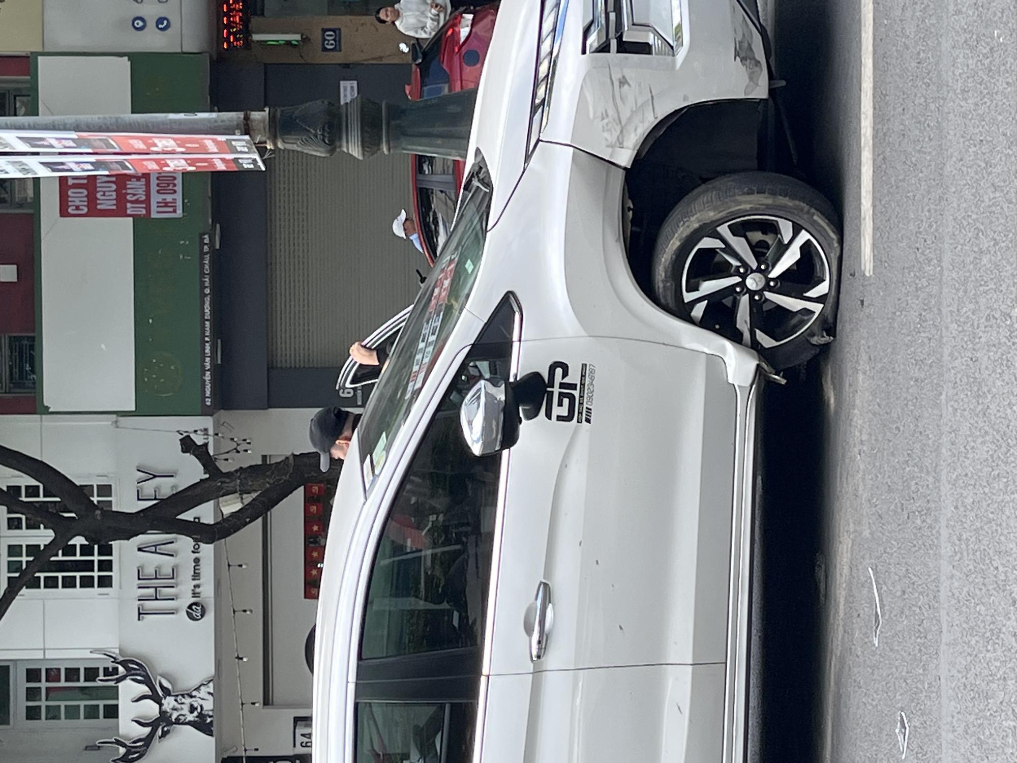 A white car, appearing to be a Mitsubishi Xpander or similar MPV/SUV, is pictured on a street in Đà Nẵng, Vietnam, exhibiting significant front-end damage. The front right side of the vehicle is severely impacted, with the bumper crumpled and partially detached, and the front right wheel turned sharply inward, indicating potential damage to the axle or suspension. The car features a "GP" logo and the phone number "0902346197" on its side.
In the background, a person wearing a black cap is partially visible near the open driver's side door of the white car. Further back, another person in a white top is seen looking into the passenger side of a red car, which is partially obscured and also appears to be involved or present at the scene.
The setting is a busy urban street during daytime with clear weather. Building facades line the street, one prominently displaying "THE ALLEY" with the slogan "@ It's time for..." and a deer logo. Another sign provides an address: "62 NGUYỄN VĂN LINH, P. NAM DƯƠNG, Q. HẢI CHÂU, TP ĐÀ", confirming the location in Đà Nẵng City. A street pole in the foreground carries an advertisement with partially visible Vietnamese text including "CHO T", "NGUY", "DT SAN:", and "LH: 0901", likely pertaining to property rental or sales. Building numbers "64" and "60" are also visible.
