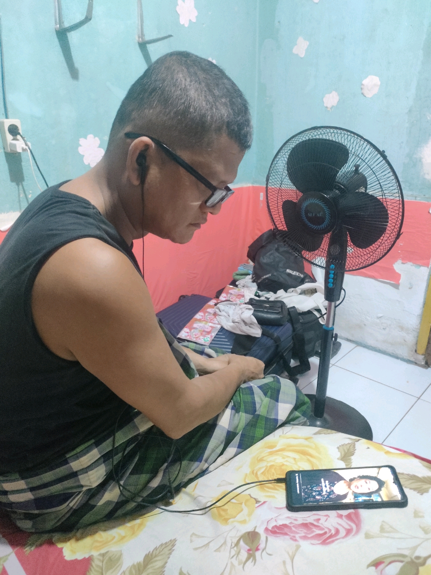 A person with short hair and glasses is sitting on a bed in Pekanbaru, Indonesia. They are wearing a black tank top and plaid pants. A black fan is standing behind them, and a smartphone is lying on the bed in front of them. The phone displays an image of a person. The room appears to be a bedroom, with a blue wall and a floral-patterned bedspread.