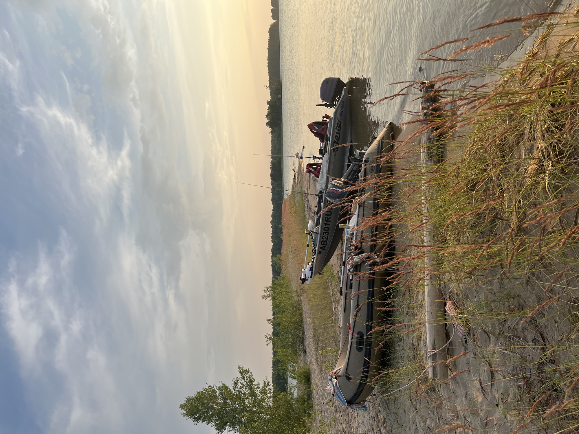 This image depicts a tranquil lakeside scene at sunset. Two inflatable boats are moored on a sandy shore, partially obscured by tall, dry grass. One boat, a tan inflatable, is positioned closer to the foreground, with a coiled rope and a piece of driftwood lying on the sand nearby. A second, larger boat, also tan with a dark motor and the registration "AB2301RU", is partially submerged in the calm, golden water, with fishing rods visible. The water reflects the soft hues of the setting sun, and the sky is a blend of pale blue and hazy white clouds. In the distance, a dense line of trees forms the horizon. The environment is natural and unpopulated, suggesting a remote or wilderness setting. The location is identified as the city of Borovoe, Russia.