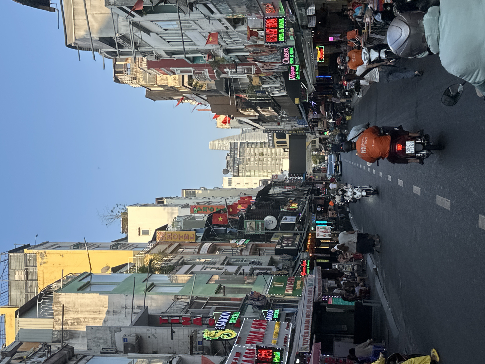 The scene depicts a bustling urban street in the city of Thủ Đức, Vietnam, under a clear blue sky, likely during late afternoon or early evening. The asphalt road, marked with dashed white lines, extends into the distance, flanked by dense rows of multi-story buildings, approximately 3 to 6 floors high. These buildings feature varied architectural styles and colors, including pale green, yellow, grey, and white, with red accents.
Numerous commercial signs, many with active neon or LED illumination in Vietnamese script, line both sides of the street, advertising businesses such as "TINA SPA," "SAIGON SPECIAL," "YUNA SPA," "KELLY SPA," "Cheers," and "Hopes." Multiple Vietnamese national flags are displayed from poles and balconies along the building facades. Overhead, a complex network of power lines and cables is visible.
Vehicular traffic includes several motorcycles and scooters in motion, some with passengers. Two individuals are operating motorcycles while wearing distinctive orange "Shopee Food" delivery uniforms. One person on a white scooter wears a face mask. Pedestrians are visible on the sidewalks and crossing the street. Groups of people are seated at outdoor tables and chairs on the sidewalks, suggesting active cafes or restaurants.
In the far background, a prominent skyscraper with a distinctive pointed apex is visible. A street sign displaying "PASTEUR STREET" is partially observable on the left side, past the mid-ground. The overall environment indicates a vibrant commercial and residential thoroughfare.
