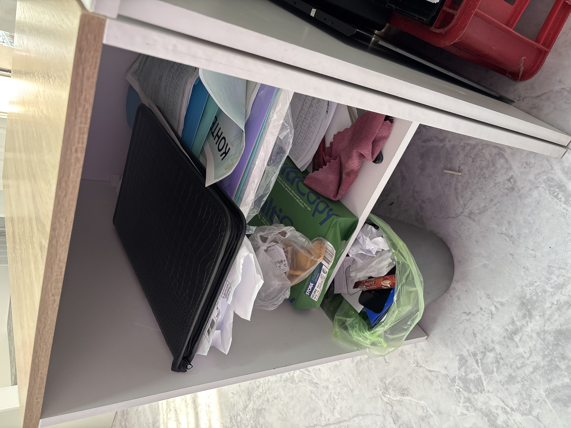 This is a top-down, eye-level shot inside a white shelf unit. The shelf is filled with various items, including folders, papers, plastic bags, and a red object in the upper right corner. A black crocodile-textured folder is on the left, with white papers sticking out. To its right are blue and light blue folders, and a plastic bag containing papers with the text "KOHTE" visible. Further to the right, a green package of paper is visible, labeled "STORA MultiCopy". A pink cloth is also present, along with a green plastic bag containing crumpled paper and a packet of "Coffee Deluxe". The shelf unit itself appears to be made of a light-colored wood on the left side and white material for the shelves. The background suggests a lightly textured, possibly marble-patterned floor. The lighting indicates an indoor setting, possibly during the daytime. No people are visible in the image.