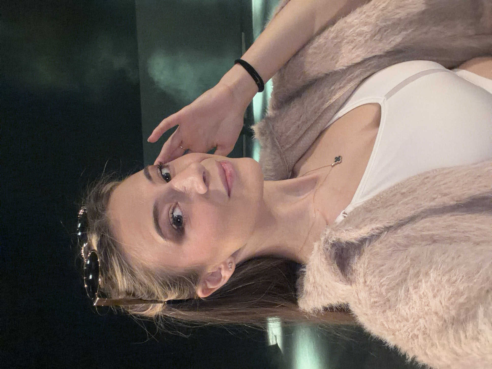 A woman is photographed indoors with dark green background. She wears a white top and a light pink fuzzy jacket. A small gold necklace is visible. She wears a black band around her wrist. A pair of sunglasses is tucked into her hair. Her right hand is near her face. The location in Plovdiv, Bulgaria is not discernible.