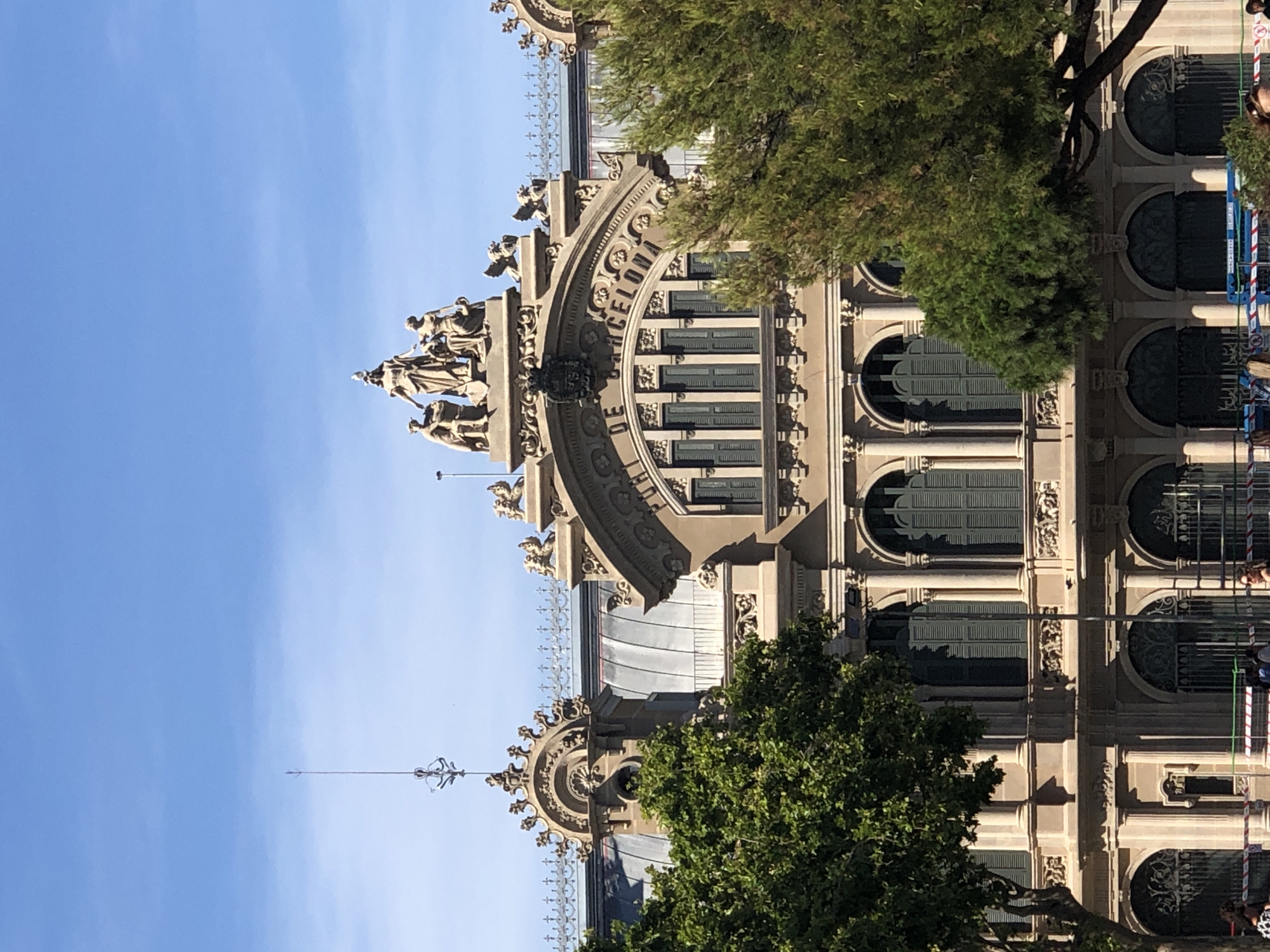 The image captures an ornate building with classical architectural features, likely a historical landmark or government building. It is topped with a sculptural group depicting figures and set against a clear blue sky. Below the sculptures, an arched pediment displays the inscription "PUNT DE BARCELONA" (Port of Barcelona). The building's facade is characterized by rows of tall windows with dark shutters, decorative friezes, and columns. Lush green trees frame the building, partially obscuring its lower sections and casting shadows. In the foreground, a glimpse of construction or event barriers with signage and people suggests ongoing activity or public access to the area. The scene appears to be a sunny day, with ample natural light illuminating the structure and its surroundings.