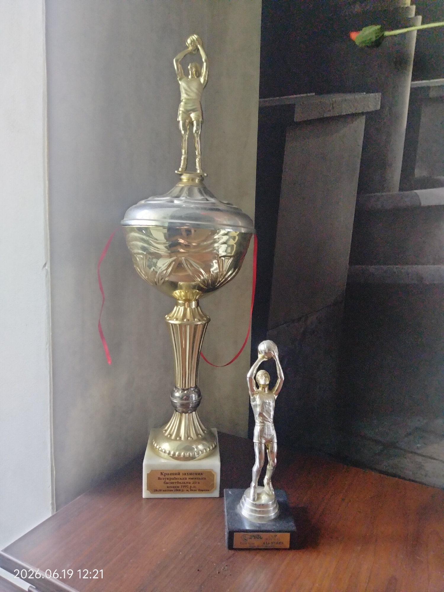 In an indoor setting, two basketball trophies are displayed on a wooden surface. The larger trophy, on the left, is silver and gold, with a large bowl and a smaller figure of a basketball player on top. A red ribbon is attached to the side of the bowl. The base of this trophy has a plaque with text in Cyrillic script. To the right, a smaller silver trophy depicts a basketball player holding a ball. This trophy sits on a black base with a plaque. The background consists of a textured wall and a dark, abstract pattern. The timestamp "2026.06.19 12:21" is visible in the bottom left corner.