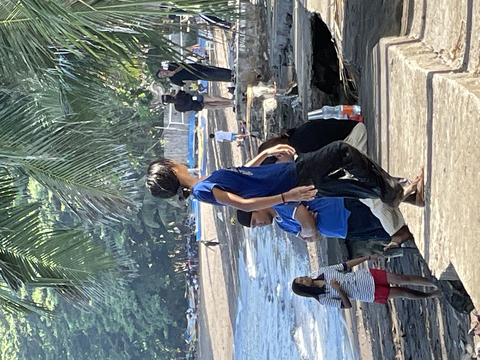 The image depicts a sunny day scene at a beach in Dauin, Philippines, likely near a coastal area with palm trees. In the foreground, a young woman wearing a striped t-shirt and red shorts walks on a concrete structure. To her right, a man in a blue shirt and black cap stands with his arms crossed, looking towards the right of the frame. Behind them, a person in a blue t-shirt and black pants walks away from the camera, their back to the viewer. Further back, several people are visible near the water's edge and on the sandy beach. The water is choppy with waves. In the background, there is dense foliage and what appears to be a building structure or pier. The overall impression is a casual gathering at a tropical beach.