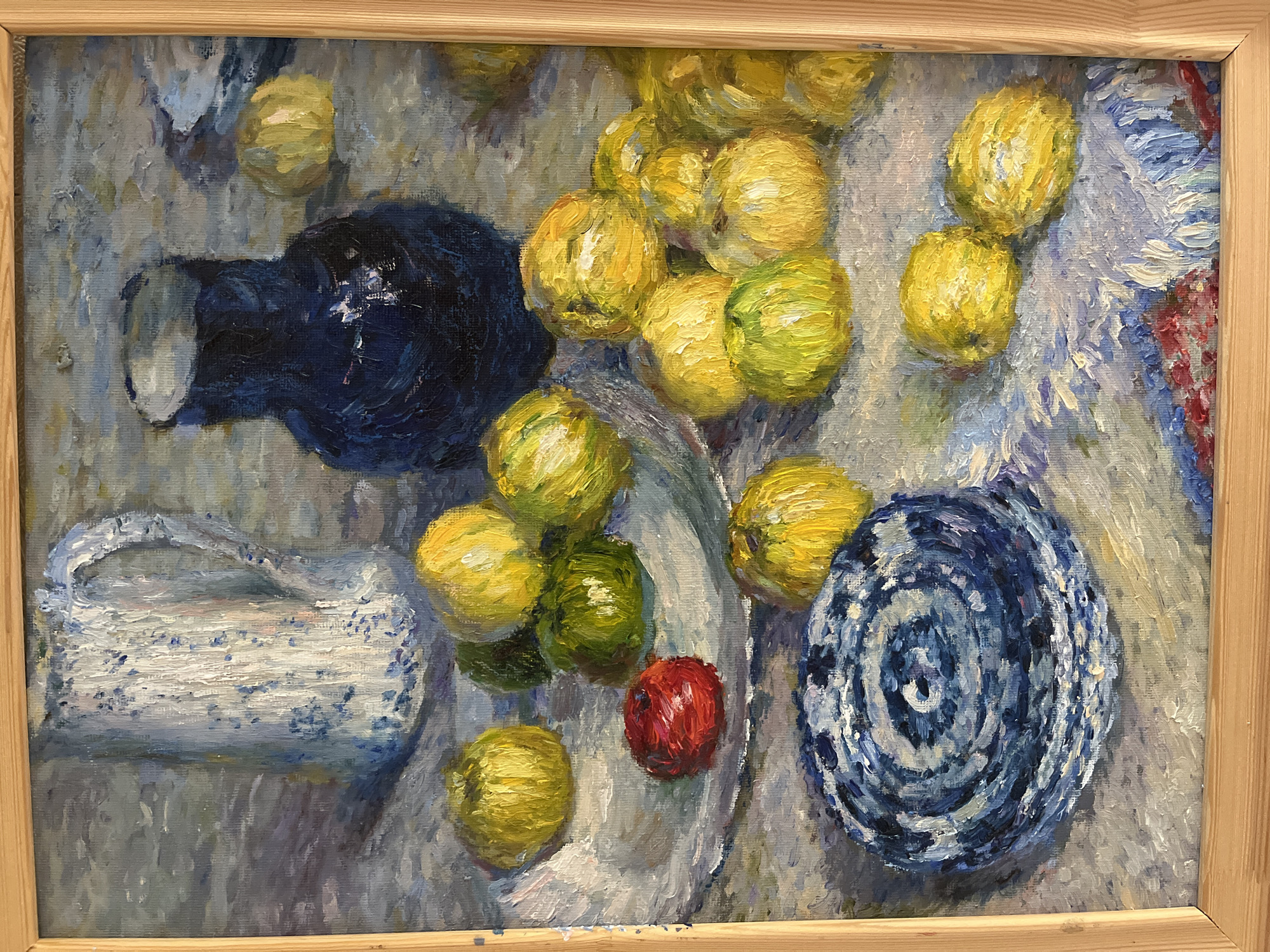 This is a framed oil painting depicting a still life. The subjects are various fruits, primarily lemons, some of which are green, and a single red apple. These fruits are arranged on a textured surface, possibly a tablecloth or draped fabric, in a loose, clustered fashion. To the left of the fruits, there is a dark blue bottle or jug lying on its side. Adjacent to the bottle is a white pitcher or jug with a blue speckled pattern and a handle. On the right side of the composition, a decorative blue and white plate or bowl with a circular pattern is visible. The overall scene appears to be set on a table, with the backdrop rendered in muted, impressionistic strokes of various grey and blue tones, suggesting an indoor setting. The lighting appears soft and diffused, typical of an interior or possibly an overcast day, though no specific time of day or weather conditions can be definitively determined. The brushwork is visible and textured, giving the painting a lively, almost tactile quality. There is no discernible text in the image. The setting is identified as Starye Politsy, Russia.