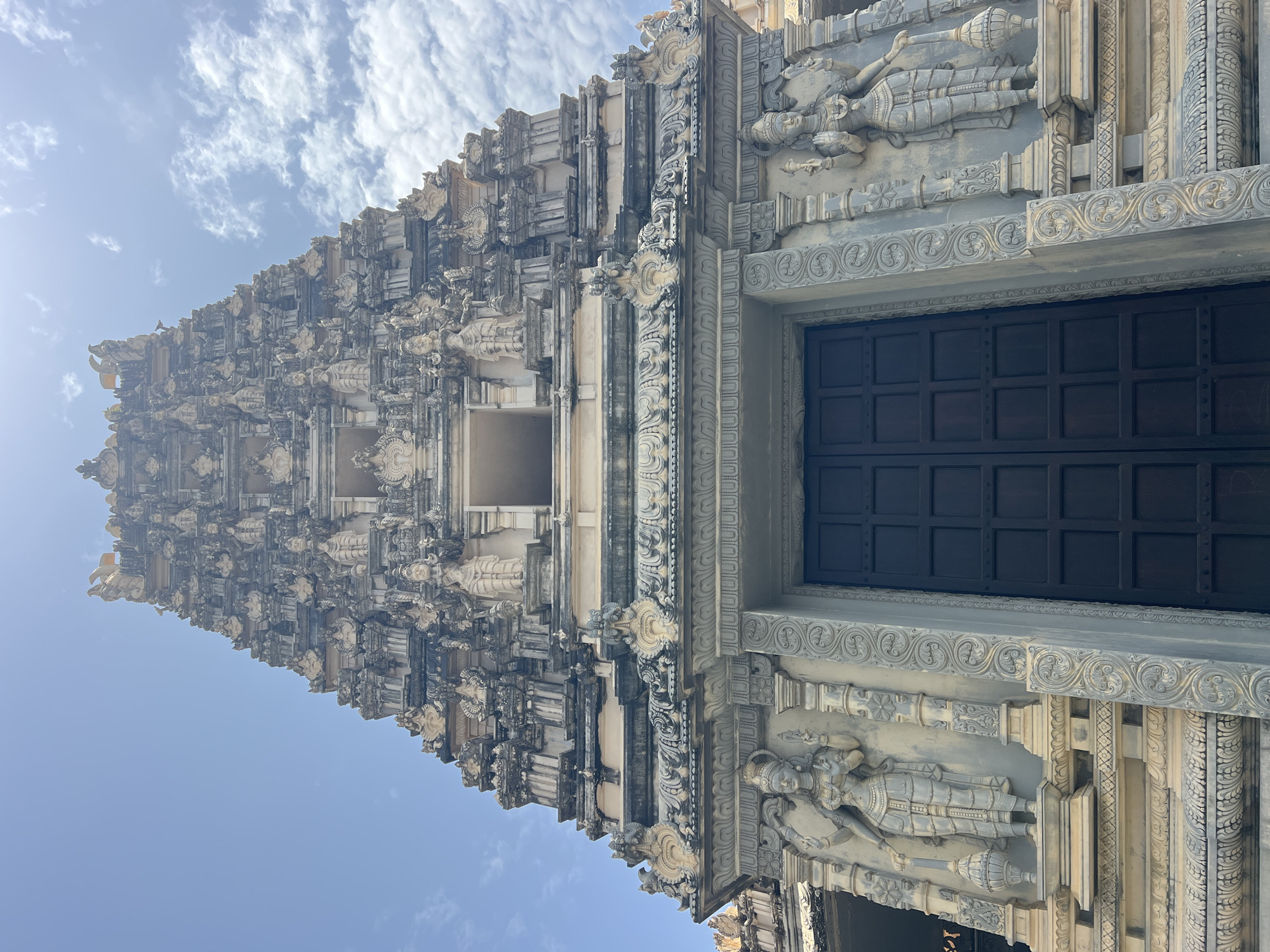 The image shows a low-angle, full-frame view of the entrance to a Hindu temple, likely the Sri Kailasanathar Swami Devasthanam Temple in Galle, Sri Lanka, given its distinctive architecture and location. The temple's towering gopuram (ornamental tower) dominates the frame, intricately carved with numerous deities, figures, and decorative motifs. The facade is constructed from weathered stone, with some sections appearing to be lighter colored sandstone or stucco, contrasted with darker grey stone elements.
At the base of the gopuram, a large, dark wooden door with a grid of square panels is centrally positioned. Flanking the doorway are two elaborately sculpted figures of female deities, each with multiple arms, standing on pedestals and adorned with traditional jewelry and attire. The architectural style is characteristic of South Indian temple construction.
The temple structure features multiple tiers of intricate carvings, including smaller figures, archways, and decorative cornices. The upper portions of the gopuram recede upwards, reaching towards a bright, partly cloudy blue sky. Sunlight illuminates the upper sections of the temple, casting subtle shadows and highlighting the detailed craftsmanship. The sky contains scattered white clouds, some of which are wispy and others more dense. The overall impression is one of grandeur and intricate artistry, suggesting a significant religious and cultural site.