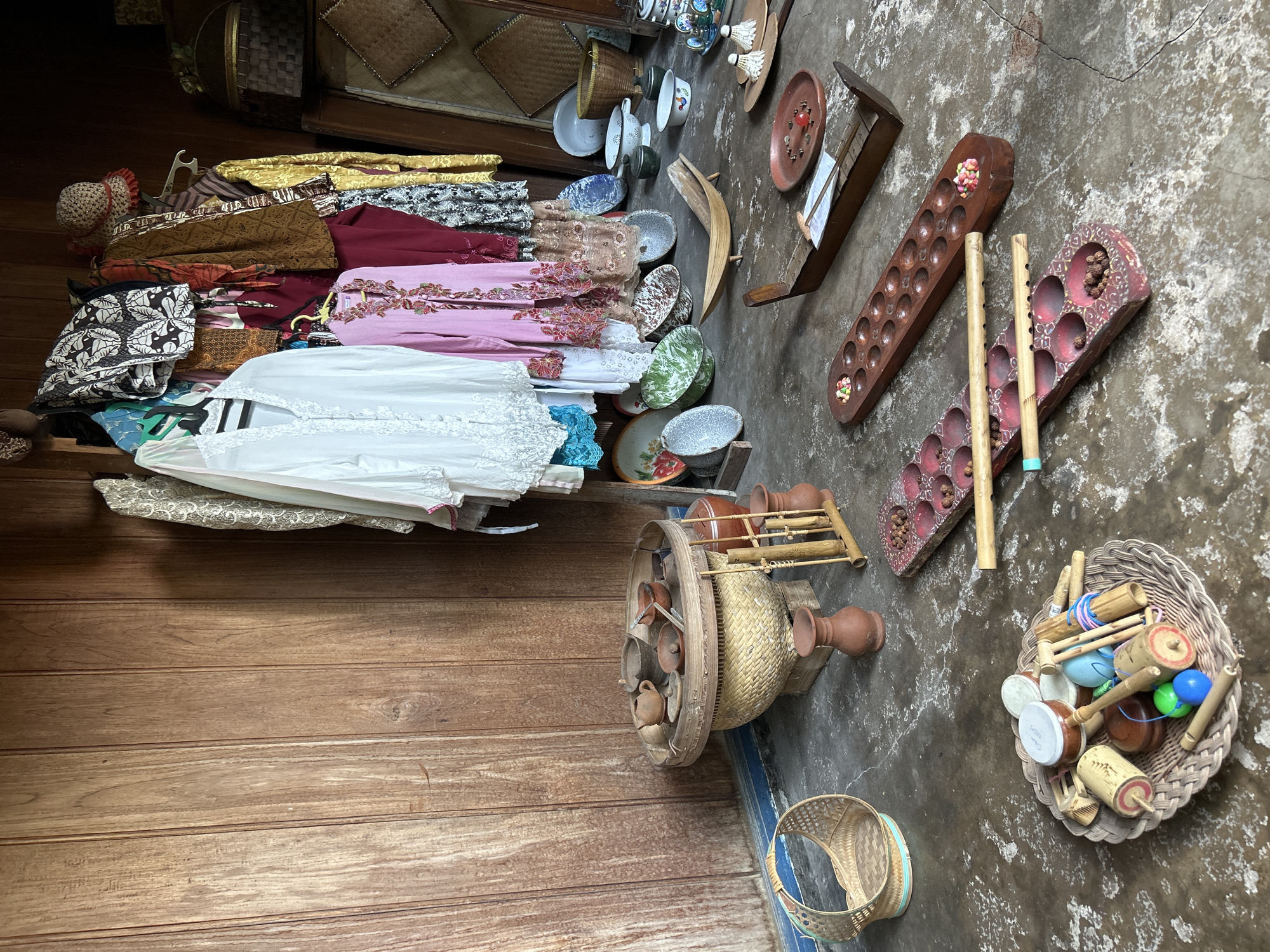 This media file presents an overhead view of a collection of traditional Indonesian items laid out on a rustic, textured floor and adjacent wooden surface. The scene appears to be a display or storage area, possibly in a shop or a home in Banguntapan, Indonesia, given the provided location context.
The objects are diverse and showcase cultural heritage. On the left, a stack of vibrantly colored fabrics, likely batik and other traditional textiles, are neatly folded. A straw hat with a red trim sits atop the fabric pile. Further right, traditional games are laid out. These include a congkak board, a wooden game with numerous holes, and other similar wooden boards with carved indentations. Scattered around these boards are small, colorful objects, possibly game pieces. Two wooden flutes also lie near the congkak board.
In the center of the frame, a large, woven basket contains a variety of small, handcrafted toys, including spinning tops, small drums, and other wooden instruments. Nearby, a round, woven tray holds an assortment of small, reddish-brown clay pots and bowls, some of which are arranged on a wooden stand.
The background reveals a concrete floor with a rough, weathered texture, and to the left, a section of wooden planks with visible grain. The overall impression is one of rich cultural artifacts, possibly awaiting sale or being preserved. The lighting suggests it might be daytime, with no specific weather conditions indicated. There is no visible text in the image.
