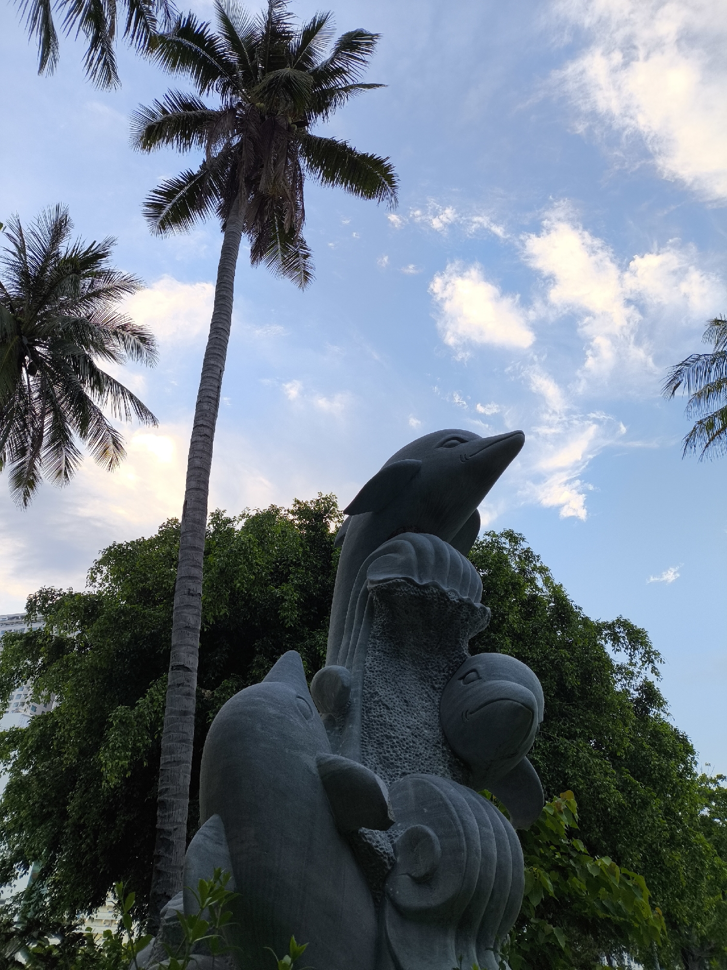 The image shows a stone sculpture of dolphins in front of a backdrop of lush green trees and a blue sky with scattered clouds. A tall, slender palm tree stands to the left of the sculpture, its fronds reaching towards the sky. In the background, a modern building can be seen peeking through the trees. The sculpture depicts several dolphins in various poses, with one prominent dolphin at the top looking upwards. The overall scene suggests a tropical or coastal environment. The location is specified as Bac Nha Trang Ward, Vietnam.