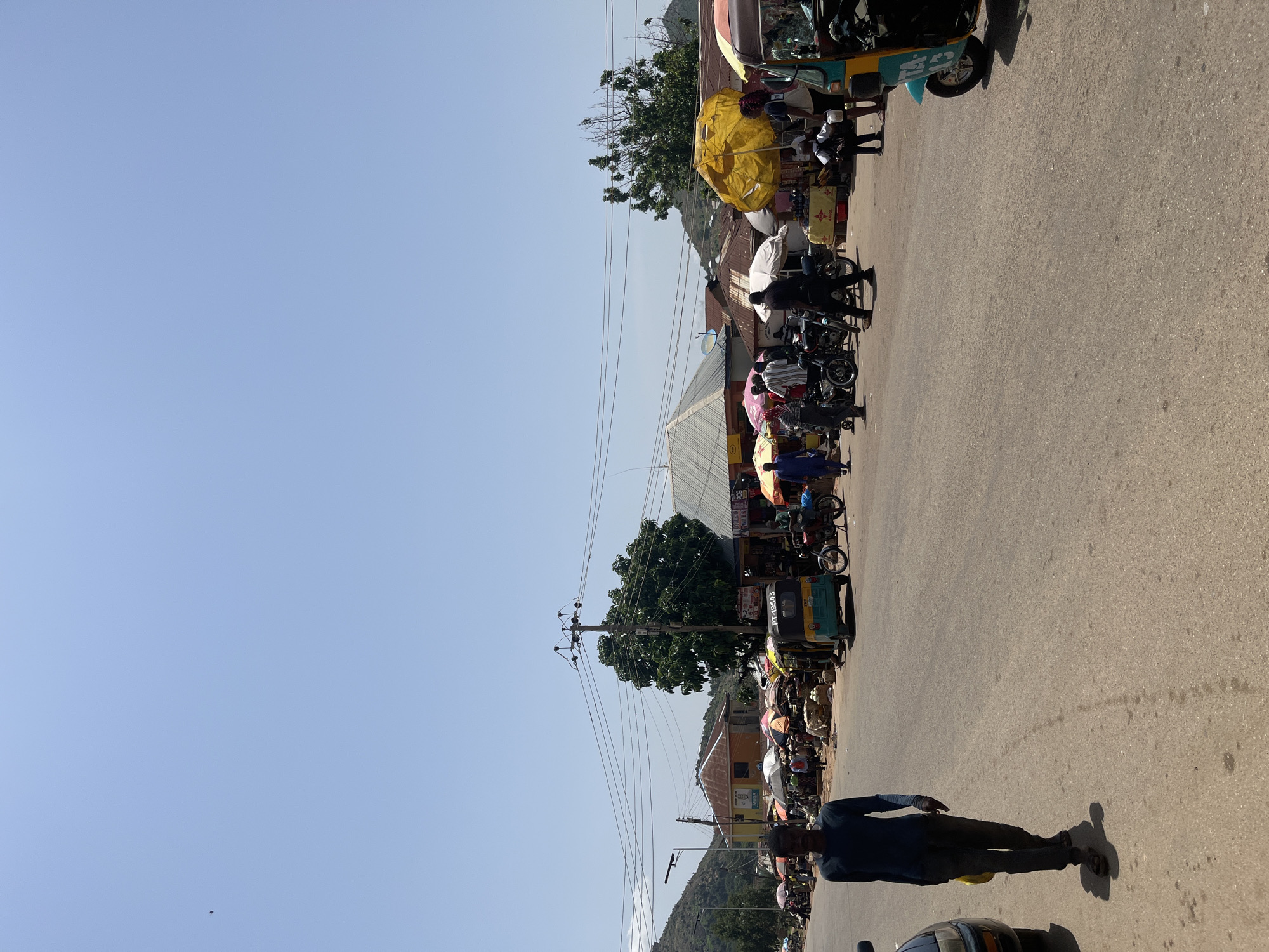 The image shows a street market scene in the city of Gwong, Nigeria. A man walks towards the camera on the left side of a paved road, wearing a dark blue long-sleeved shirt and dark pants. Behind him, a dark colored car is partially visible on the left edge of the frame. The road leads into a busy market area with numerous stalls and vendors under colorful umbrellas. Several people are browsing or working at the stalls. Motorcycles and auto-rickshaws (also known as Keke Napep) are present, with one auto-rickshaw in teal and yellow parked on the right side of the road. In the background, buildings with corrugated metal roofs and a large tree are visible. Hills can be seen in the distance under a clear blue sky. The scene is illuminated by bright daylight, suggesting it is mid-day.