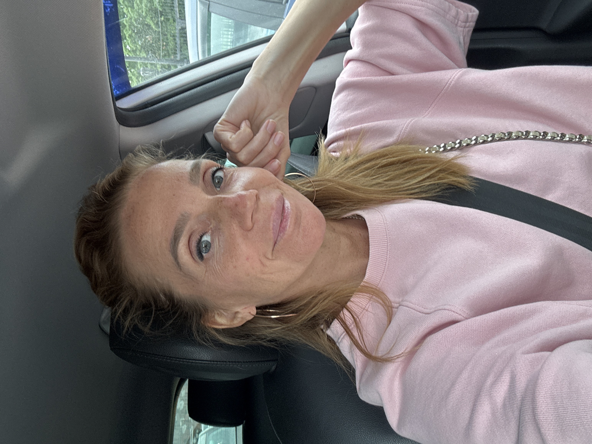A blonde woman with light blue eyes and freckles is sitting in the driver's seat of a car. She is wearing a light pink crewneck sweatshirt and large gold hoop earrings. Her left arm is resting on her chin, and she is looking directly at the camera. The interior of the car is visible behind her, with a black leather seat and the ceiling. Outside the car window, trees and other vehicles can be seen. The image appears to be a selfie taken during the daytime. The specific location in Saint Petersburg, Russia cannot be determined from the image alone.