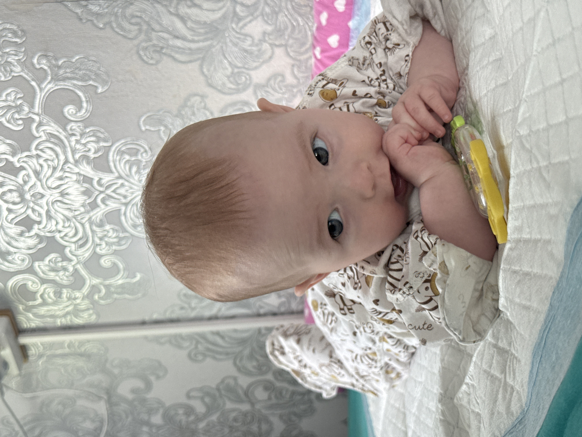 The image shows a baby lying on a white quilted surface with a light blue edge. The baby is wearing a white patterned onesie. A yellow and clear plastic object is near the baby's hands. The background includes a wall with a gray and white floral pattern and a glimpse of a pink surface with white hearts. The image was taken in Кемуль, Russia.