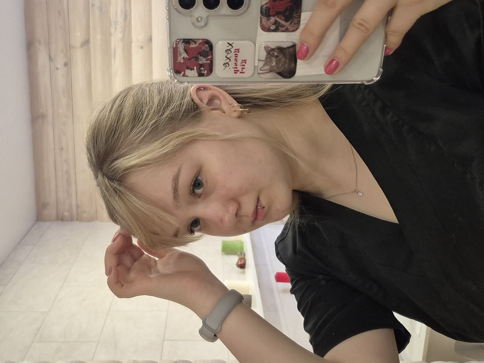A woman is taking a selfie in a bathroom. She has blonde hair and blue eyes, and is wearing a black shirt and a gray watch. Her nails are painted pink. She is holding a cell phone with a clear case and several stickers on the back. The bathroom has white tile floors and light wood paneled walls. In the background, there are various bathroom items, including a red object and a green object. The location of the bathroom is likely in Chaikovsky, Russia.