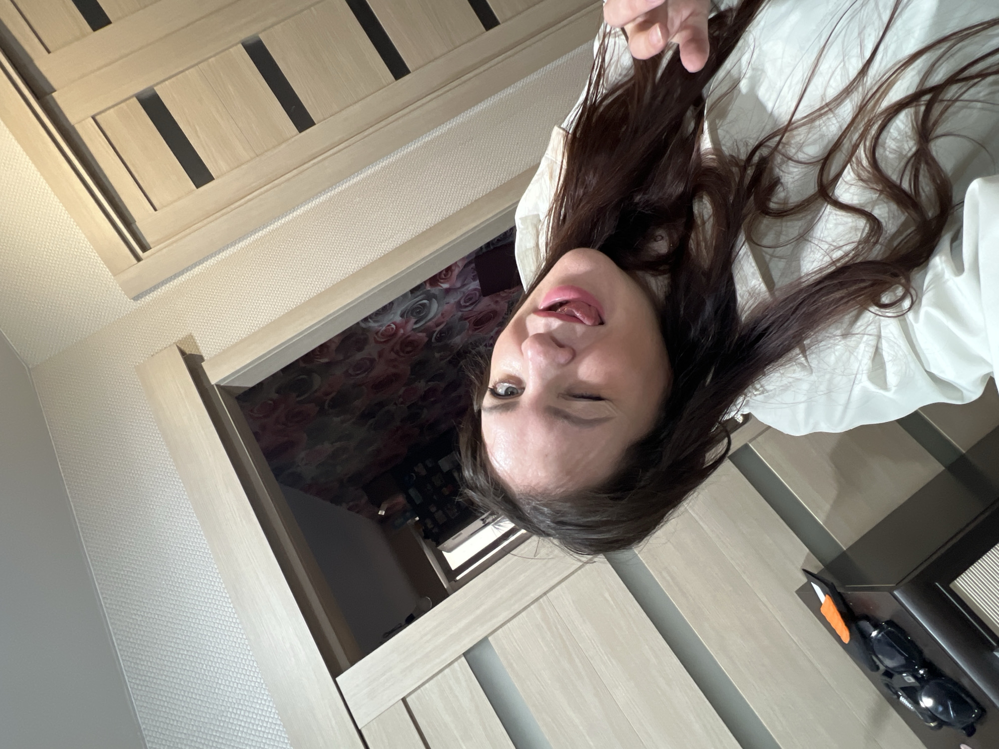 A playful selfie captures a woman indoors, making a winking face and sticking out her tongue. Her long, dark brown hair frames her face, and she appears to be wearing a light-colored top or shirt.
The background shows an interior setting, likely a room within a home. To the left (when viewed upright), there are modern-looking light wooden doors or panels with dark horizontal accents, possibly a wardrobe or closet. Adjacent to these doors, a textured, light-colored wallpaper or wall covering is visible. Through an open doorway or reflection in the background, another room is partially visible, featuring dark wallpaper with a prominent floral pattern, possibly red roses.
To the right of the woman, a dark surface, perhaps a nightstand or shelf, holds a pair of dark sunglasses and a small orange object. Further in the distance, a bright light suggests a window, indicating it is daytime. The overall mood is casual and informal, captured in a personal moment. The scene is consistent with an indoor setting in Noviy, Russia. No visible text is present in the image.