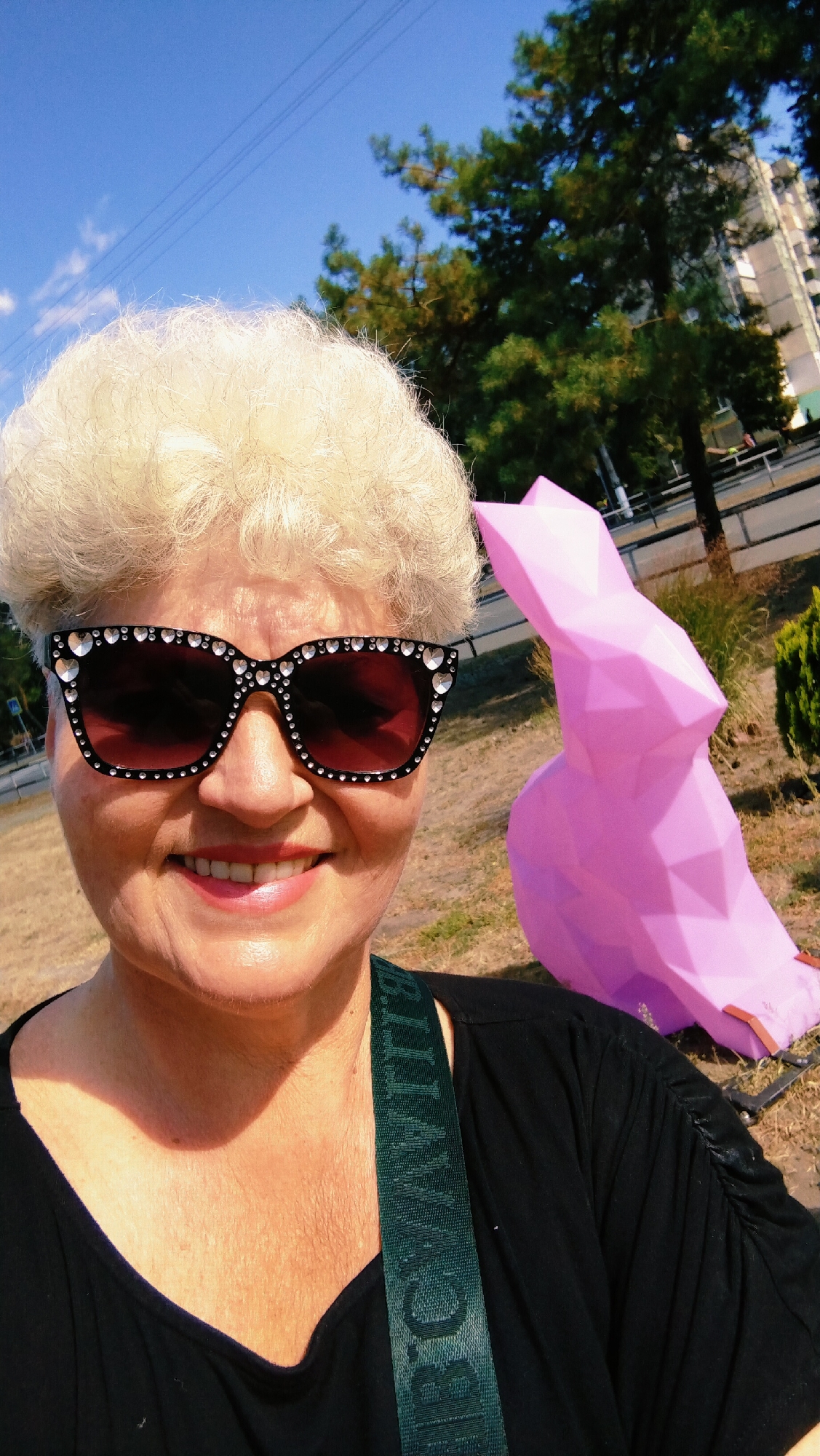 A woman with blonde, curly hair and sunglasses adorned with rhinestones smiles at the camera. She is wearing a black shirt and a green strap with text is visible across her chest. Behind her, a large, pink, polygonal sculpture of a rabbit is situated outdoors. The background includes dry grass, trees, a building, and a clear blue sky with power lines. The location is identified as Horishni Plavni, Ukraine.