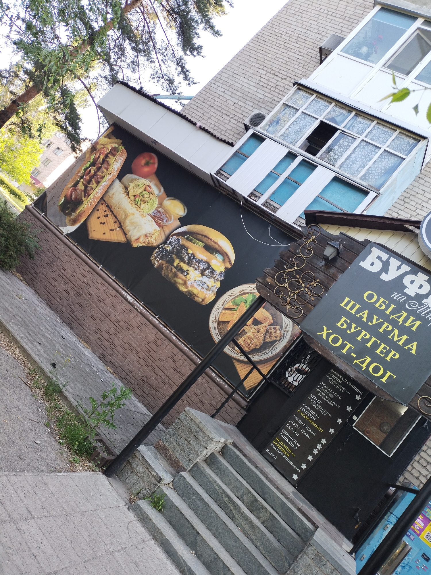 The image shows the exterior of a fast-food establishment, likely a cafe or diner, located in the city of Horishni Plavni, Ukraine. The facade features a large advertisement mural depicting a variety of fast food items, including a large hot dog, a burrito, and a burger. The establishment has a prominent sign with Cyrillic lettering that appears to list menu items such as "ОБІДИ" (Lunches), "ШАУРМА" (Shawarma), "БУРГЕР" (Burger), and "ХОТ-ДОГ" (Hot Dog). There is also a smaller menu board with more detailed offerings in Cyrillic. The building itself has a tiled roof and some windows with decorative curtains. A black lamppost stands in front of the establishment, and a set of concrete stairs leads up to an entrance. The area surrounding the building includes a paved sidewalk, some greenery, and a brick wall. The sky is visible at the top of the image, with some trees. The overall scene depicts a typical urban commercial setting.