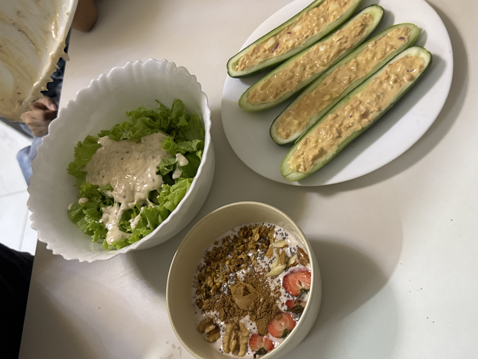 In a well-lit indoor environment, possibly a home kitchen, three dishes are presented on a neutral-colored countertop. To the left, a light brown bowl contains a breakfast or snack item, likely yogurt or a smoothie base, topped with granola, chia seeds, sliced strawberries, almonds, and walnuts, with a dusting of cocoa powder. In the center, a white, scalloped-edge bowl holds a fresh green lettuce salad, partially covered with a creamy, speckled dressing being poured from above by an unseen hand. To the right, a white oval plate displays four halves of cucumber, hollowed out and filled with a light brown mixture, possibly a tuna or egg salad. The scene appears to be a home setting, with a suggestion of movement from the dressing being poured. There is no discernible connection to Davao City, Philippines.