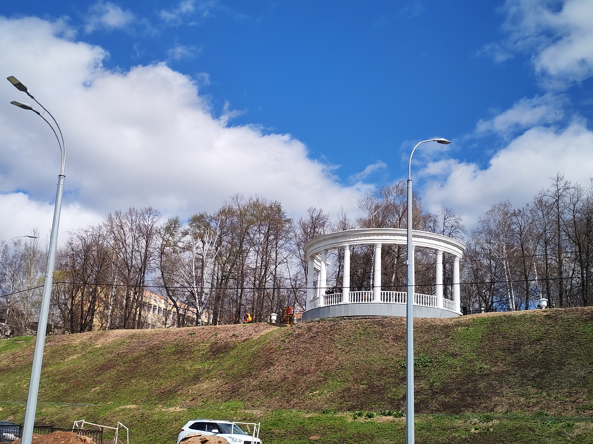 A daytime shot in Izhevsk, Russia, shows a white gazebo with columns atop a grassy hill, framed by trees. Two silver streetlights stand on either side of the hill. A white SUV is parked at the bottom of the hill near a construction site with orange safety fencing. The sky is blue with scattered white clouds. There are a couple of people standing inside the gazebo.