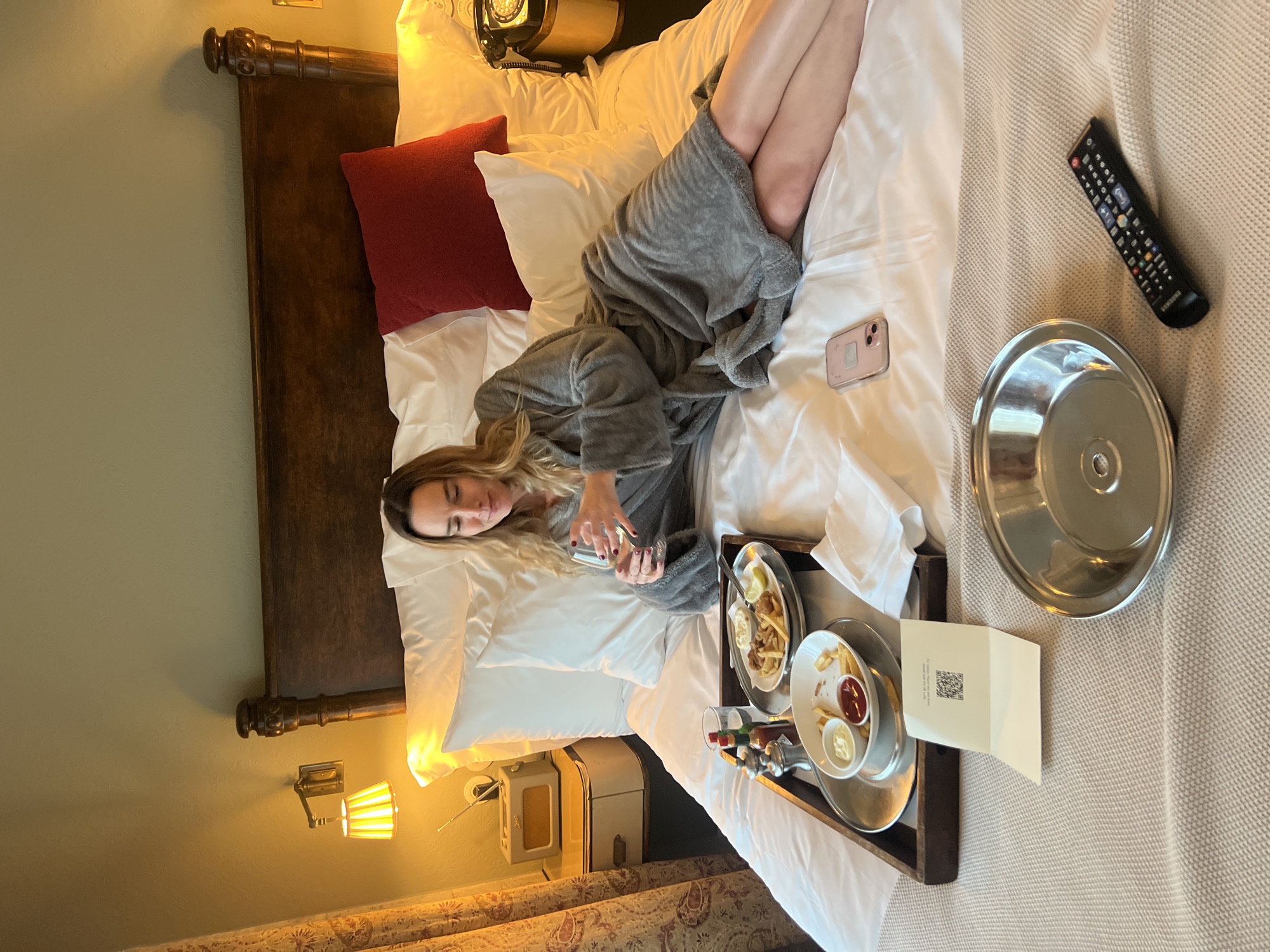 A woman in a grey robe reclines on a bed with white linens in an interior space. A wooden headboard is visible at the top left, and bedside tables flank the bed. A red throw pillow sits near the headboard. A tray of food, including plates of what appears to be fries, condiments, and drinks, rests next to her on the bed. A smartphone, a television remote control, and a metal dish are on the bed. The scene appears to be indoors, possibly a hotel room. No specific location in Berlin, Germany, can be determined from the image alone.