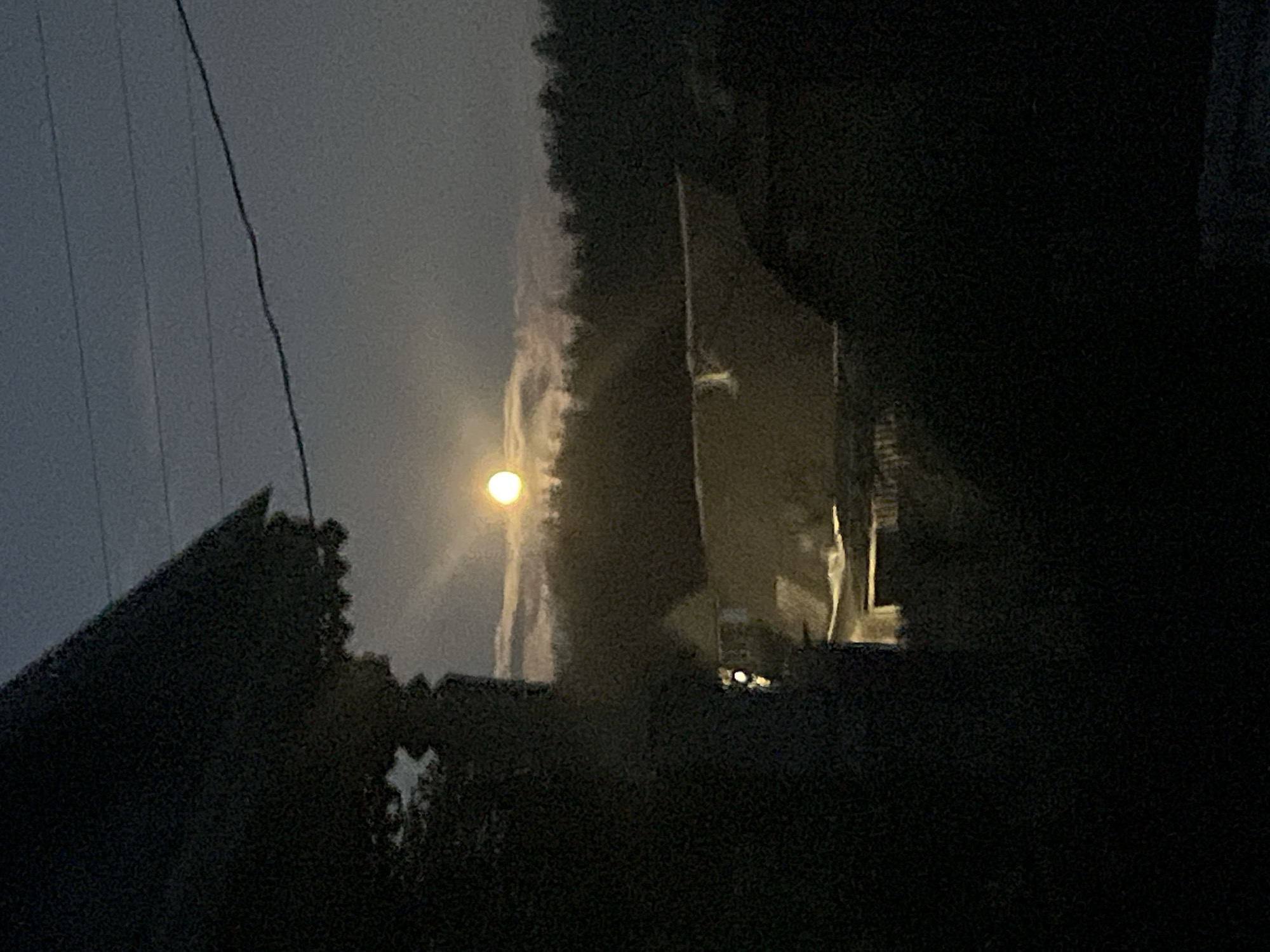 An outdoor nighttime shot shows a full moon in a cloudy sky over a darkened landscape. In the foreground, the edge of a dark structure, likely a building or a large tree, partially obscures the view on the left. Below this, another structure with illuminated windows is visible, suggesting a residential area. In the background, a dense line of trees forms a dark silhouette against the sky. The scene appears to be a rural or suburban area with a modest dwelling. The overall impression is one of quiet and stillness under the moonlight. The location is specified as Nizhny Tagil, Russia, a city known for its industrial heritage and surrounding natural beauty.