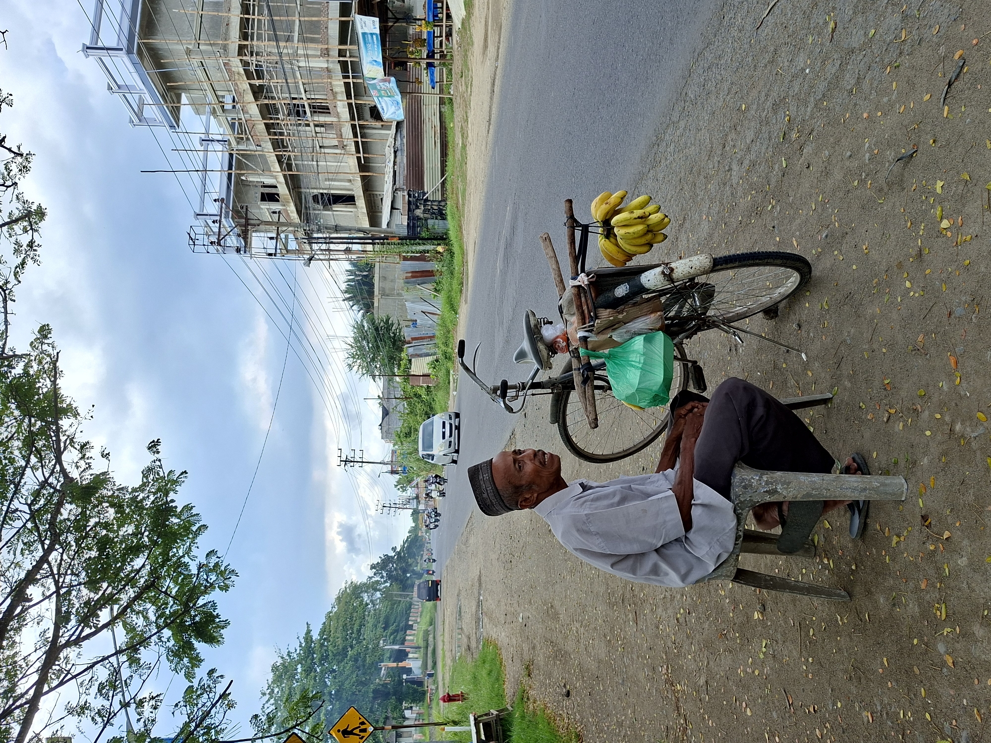 An elderly man sits on a metal stool next to a bicycle loaded with bananas on the roadside in Lhokseumawe, Indonesia. The man is wearing a light-colored shirt, dark pants, and a taqiyah (Islamic skullcap). The bicycle is loaded with a bunch of bananas hanging from the handlebars. A green plastic bag is visible on the bicycle as well. Behind the man is a road with light vehicular traffic. Further back is a building under construction. Trees and power lines are visible in the background. The ground is a mix of asphalt and dirt covered with fallen leaves. The sky is partly cloudy.