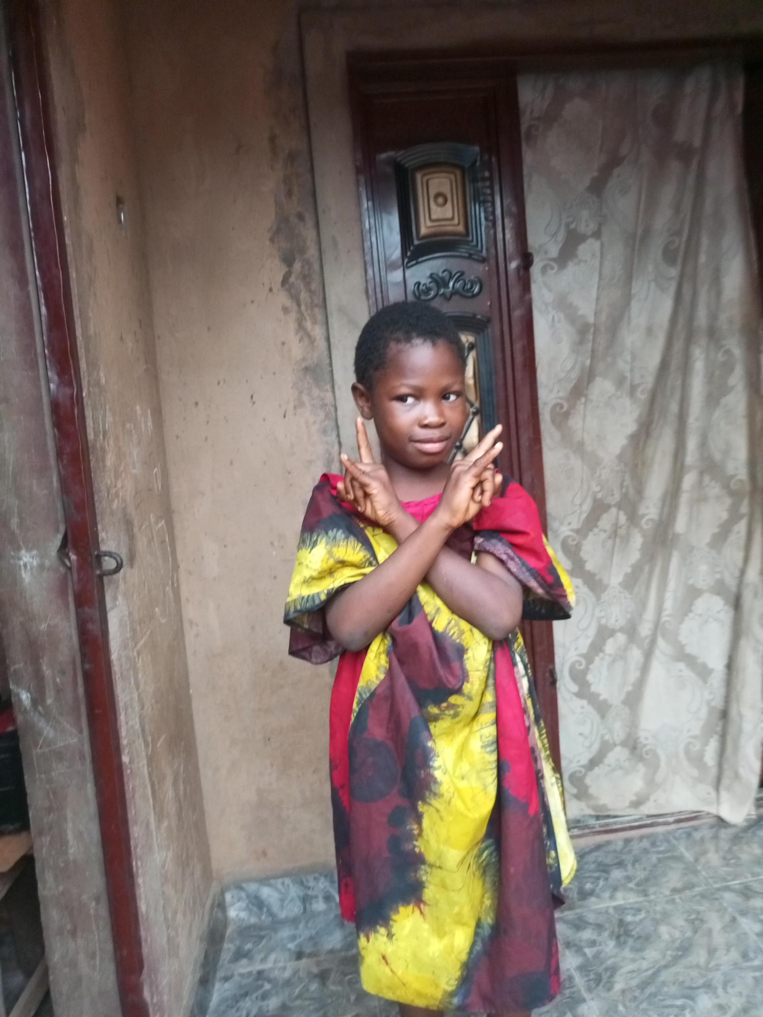 A young Black girl, appearing to be around 8-10 years old, stands in what appears to be an outdoor area in front of a building. She is wearing a colorful dress with a pattern of red, yellow, and black abstract shapes. The girl is looking slightly to her left, with her arms crossed in front of her chest and her index fingers extended upwards in a peace sign. The environment suggests a residential area, possibly in Tempure, Nigeria. To her left is a wall that appears to be made of concrete or stucco, with some graffiti or markings. To her right, there is a wooden door with ornate detailing, and behind that, a patterned curtain. The ground is covered in grey tiles. The lighting suggests it is daytime.