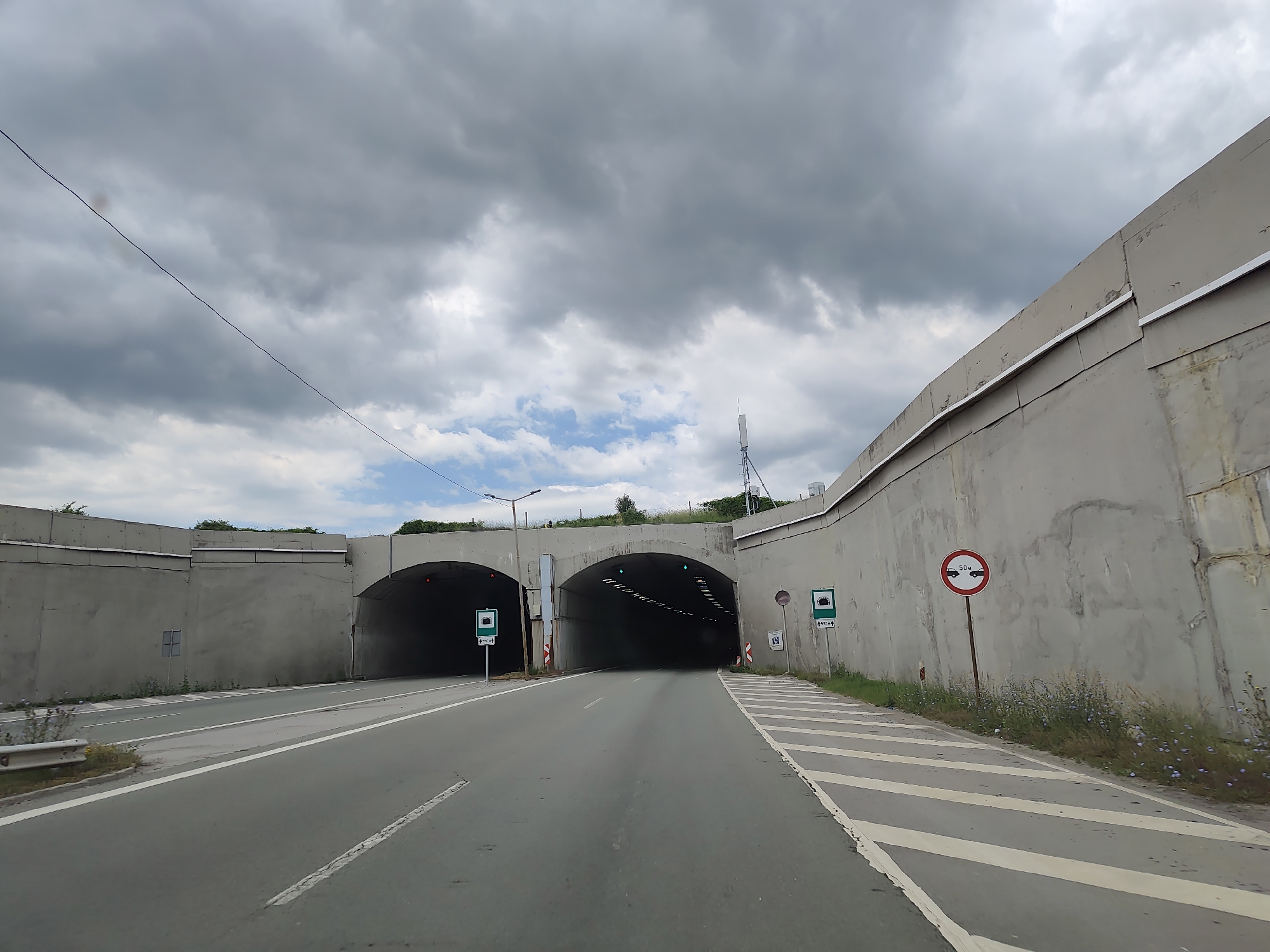 The image shows a view of two adjacent tunnel entrances on a road in Golemo Buchino, Bulgaria. The road leading to the tunnels is paved with asphalt and has white lane markings. On either side of the road are concrete walls and embankments.
There are several traffic signs visible. On the left side, a green sign with a symbol of a tunnel is present. On the right side, a circular red and white sign indicates a speed limit of 50 km/h, with a symbol of a truck. Another green tunnel sign is also present on the right.
The sky is overcast with dark clouds, suggesting a possibility of rain. Some vegetation, including grass and small flowering plants, is visible along the sides of the road and the embankments. A metal guardrail is also present on the left side of the road. The tunnels themselves are dark, with visible lighting inside, and green lights indicating they are open. A telecommunication tower is visible in the background on the right side. A thin wire stretches diagonally across the sky from the left to the center.