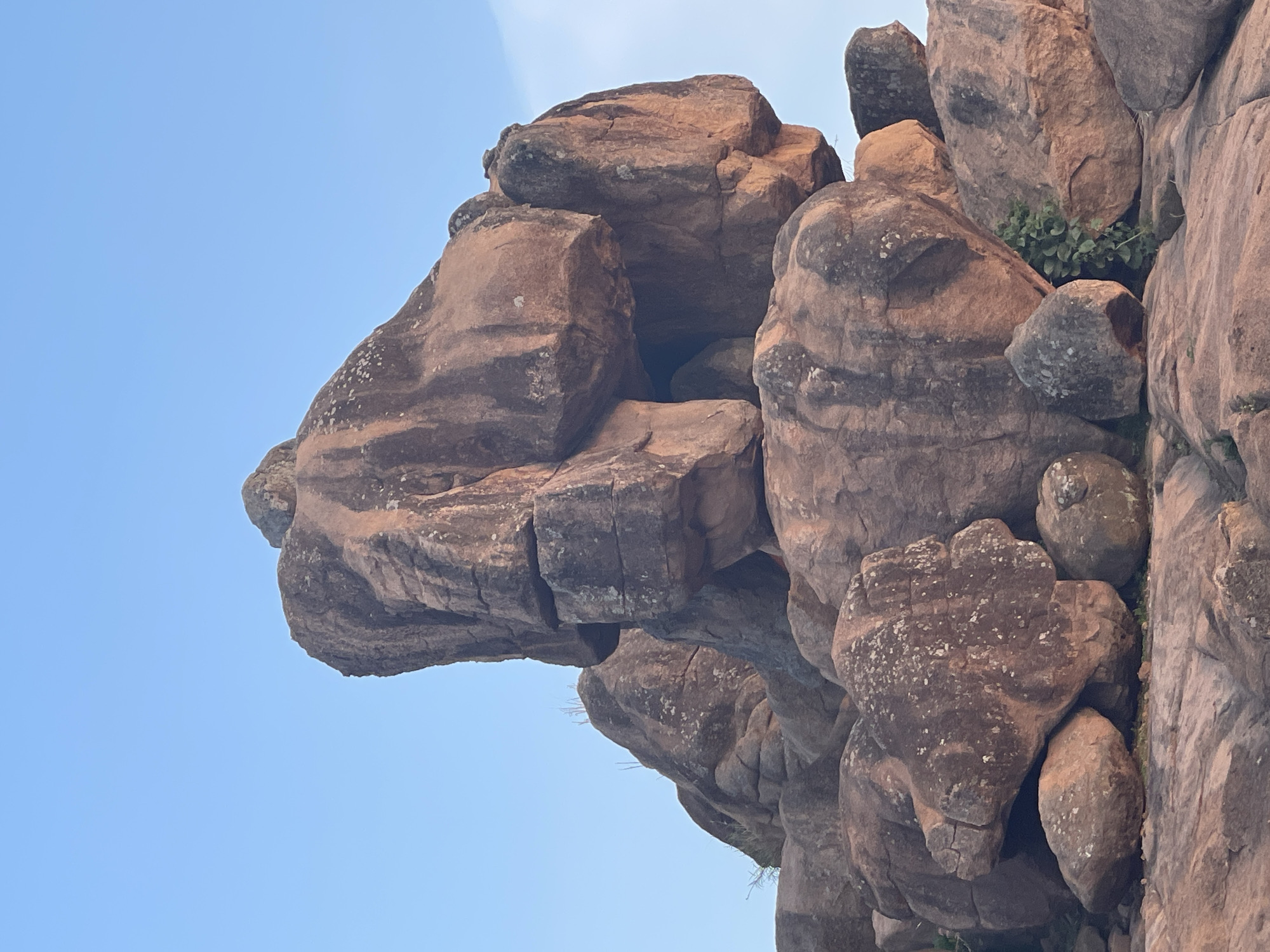 A close-up, rotated shot shows a rock formation in Barikin Gidar Kanat, Nigeria. The rocks are predominantly brown with patches of grey and some green vegetation growing on the side. The sky is light blue in the background. The photograph is taken at an angle, making the rocks appear to tower against the sky. The texture of the rocks is rough and weathered.