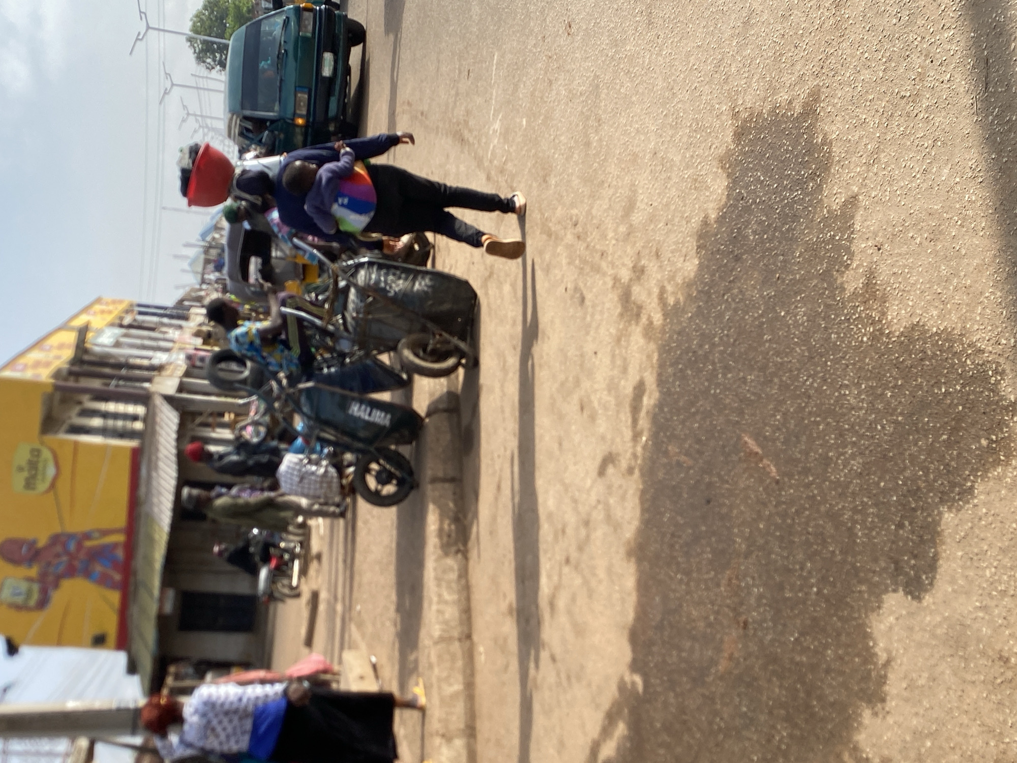The image shows a street scene in Bukuru, Nigeria. A group of people are visible near a row of motorcycles and a vehicle. A person is walking on the street carrying a red bucket and a colorful bag. The building has a yellow sign with a red figure on it.
