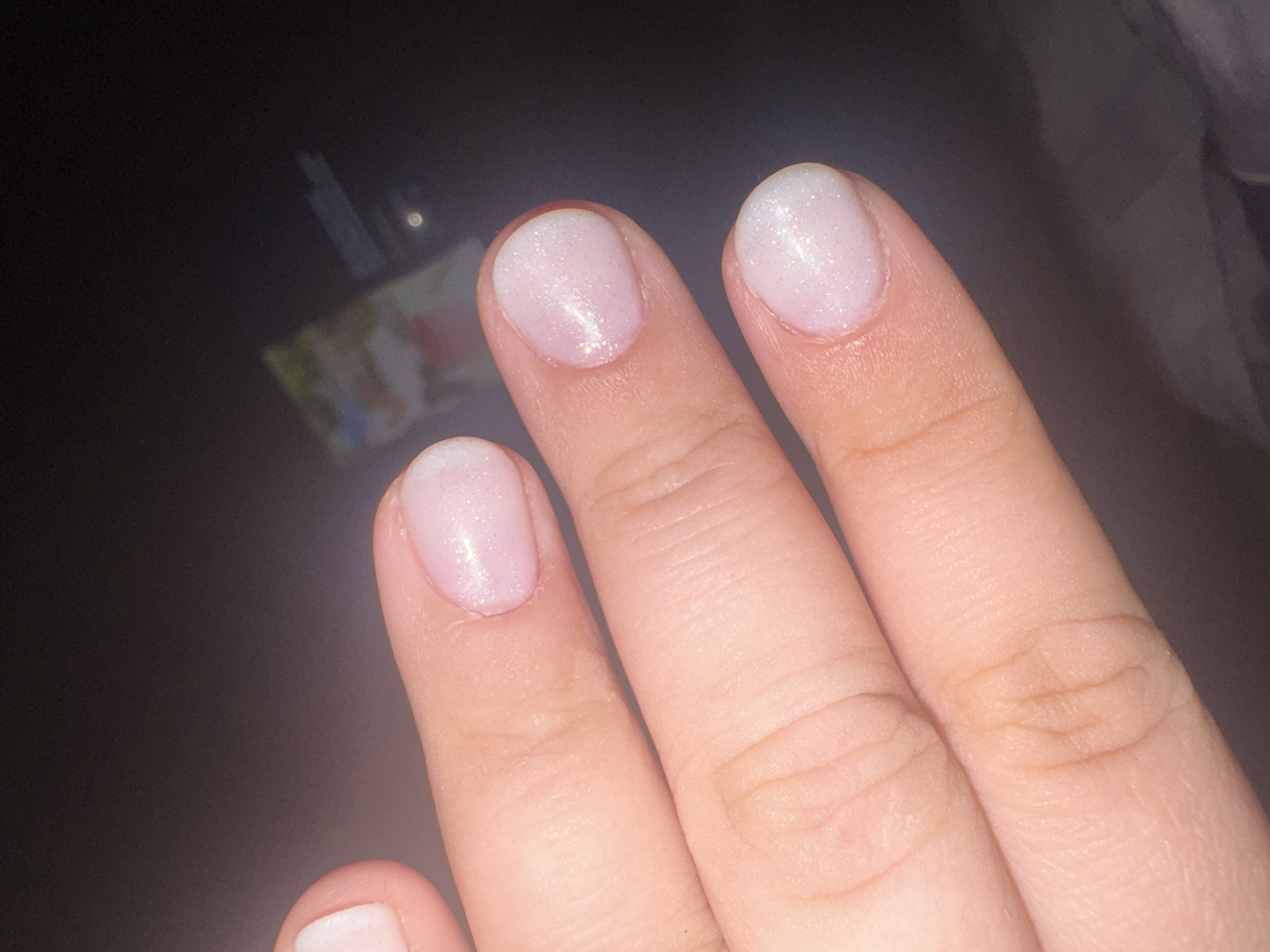 The image shows a close-up view of a person's fingers with light pink, shimmery nail polish. The focus is on the nails and the skin of the fingers, which appear healthy and well-manicured. The background is dark and out of focus, suggesting the photo was taken in a dimly lit environment. There is no indication of the location within the city of Saratov, Russia, or any specific environmental details that would place it there. The image is purely a portrait of hands and nails.