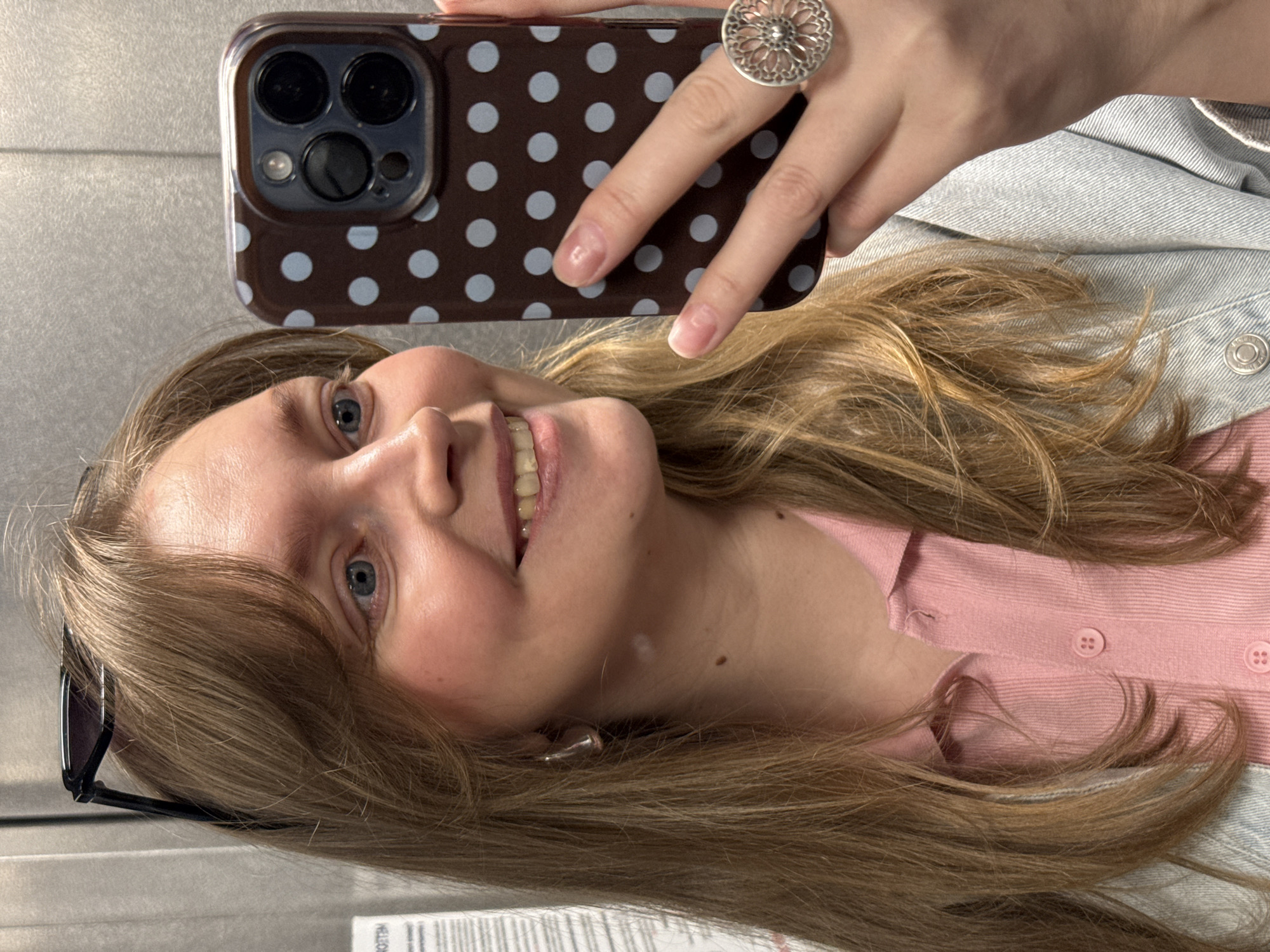 A person is taking a selfie with a smartphone encased in a brown case with light blue polka dots. The phone is held in the upper center of the frame by a hand adorned with a silver ring that features a floral design. The person is wearing a light pink top, layered under a light blue denim jacket. A pair of sunglasses is positioned near their face. The person has light hair and blue eyes. The background is a gray, textured surface. There is no indication that the photo was taken in Izhevsk, Russia.