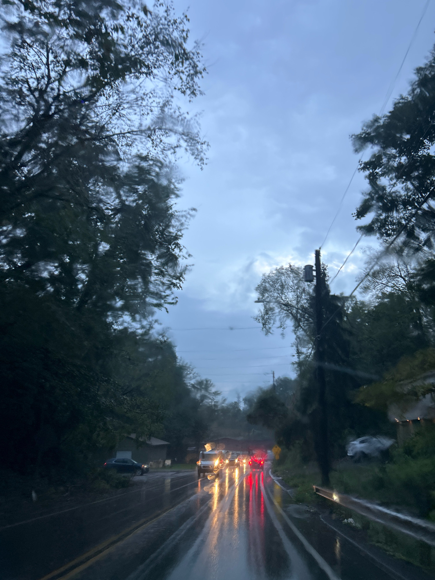 The image depicts a rainy scene on a road, likely a residential street in Scott Township, United States of America. The perspective is from inside a vehicle, evidenced by raindrops on the windshield and the view of the road ahead. Several vehicles are visible, their headlights and taillights reflecting on the wet asphalt, creating streaks of light. Trees line both sides of the road, their leaves appearing dark and wet. A utility pole with electrical equipment stands on the right side of the road. The sky is overcast with clouds, suggesting inclement weather. The overall impression is one of a damp, somber atmosphere, characteristic of a rainy day.