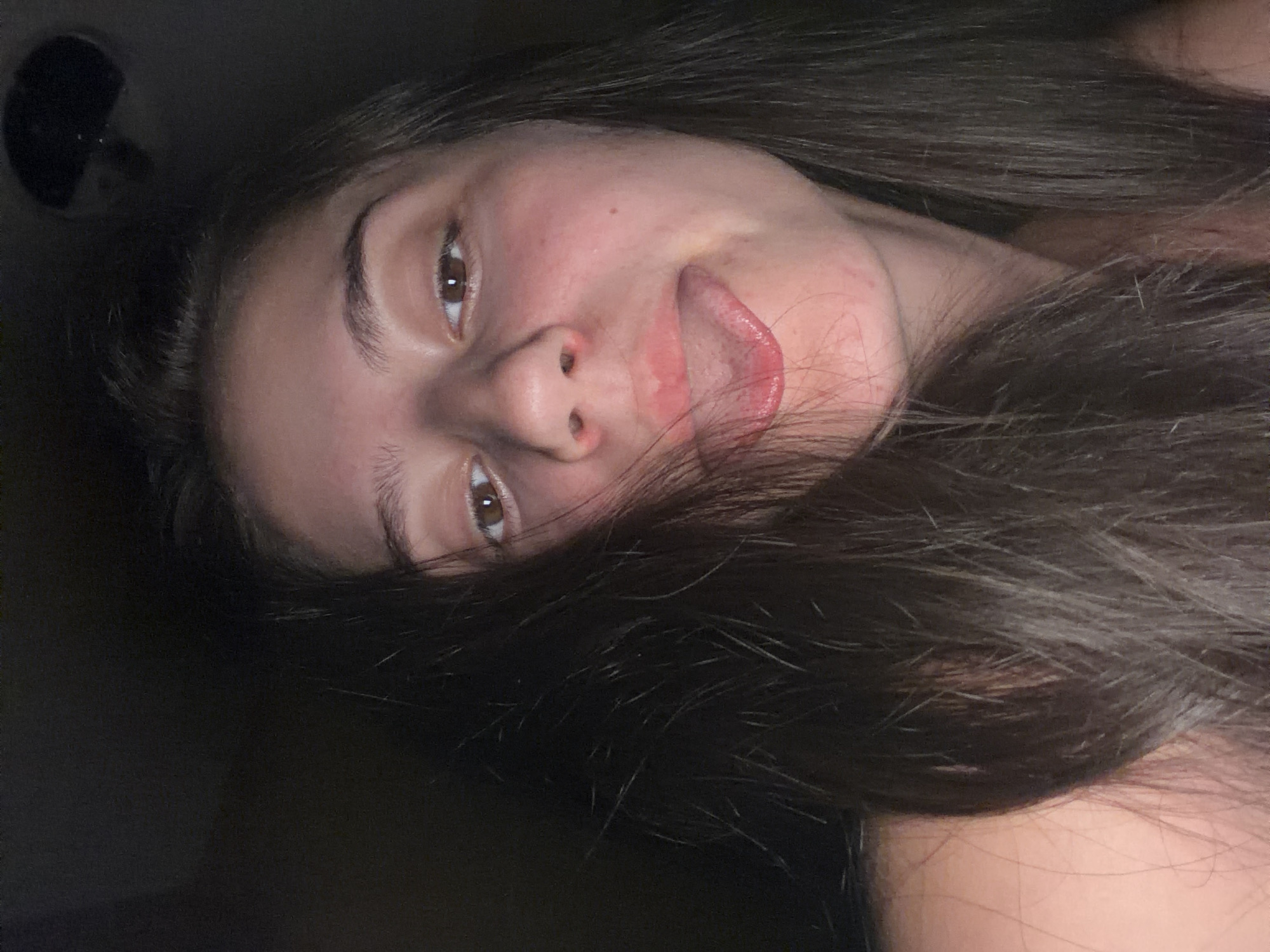 The image is a close-up selfie of a young person with long, dark hair. The person is looking at the camera with their tongue sticking out and slightly to the side. Their skin is fair with some redness on the cheeks. The background is dark and out of focus, making it impossible to determine the location in Burgas, Bulgaria, or any specific environmental details.