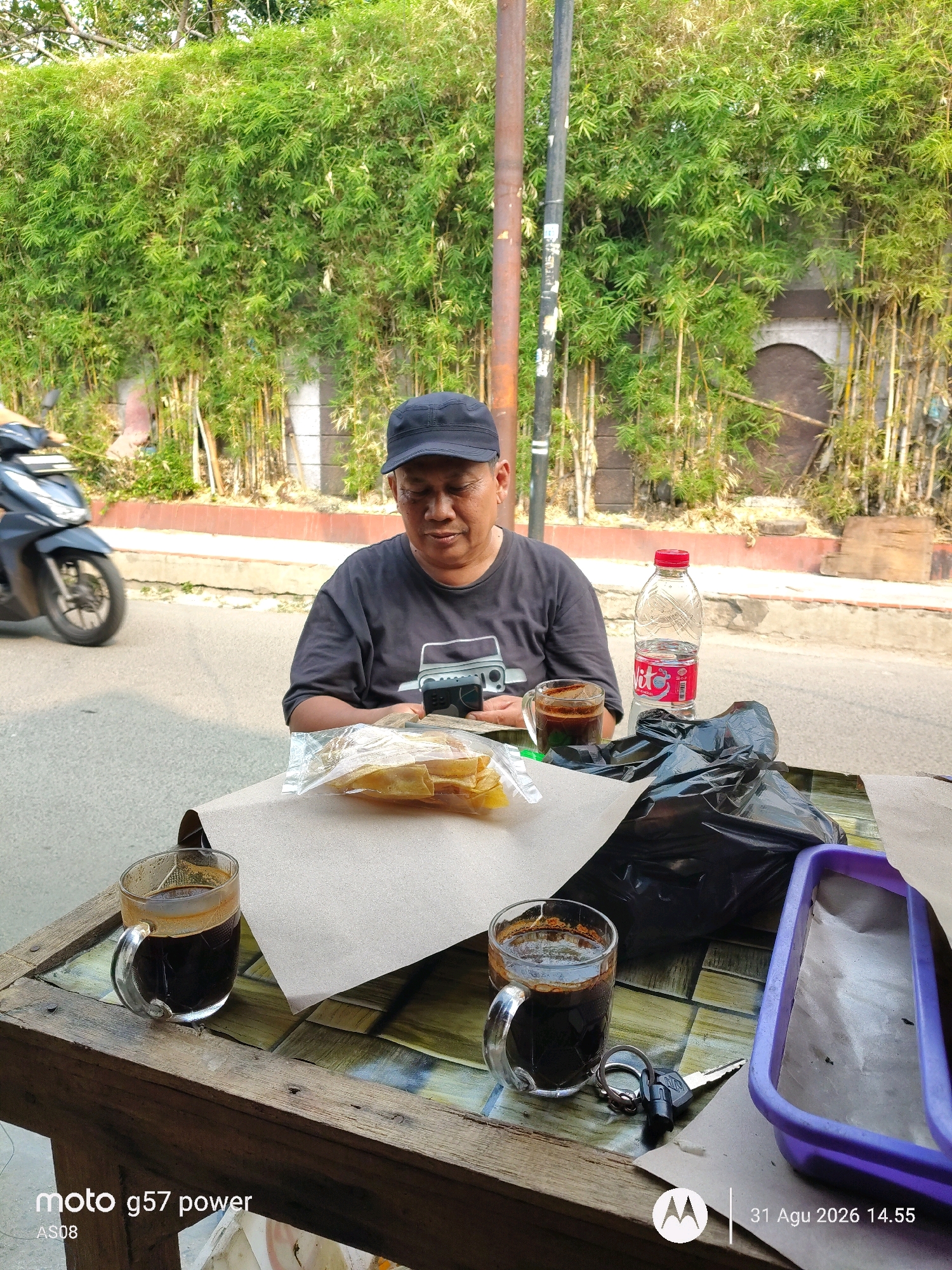 A man wearing a black cap and t-shirt sits at a wooden table outdoors, looking down at his phone. In front of him are two glasses of coffee, a bottle of red-capped soda, a plastic bag filled with food, and a set of keys. A motorcycle is visible in the background, as is a wall of bamboo plants. The scene appears to be on a street in South Jakarta, Indonesia, based on the "moto g57 power AS08 31 Agu 2026 14.55" text overlay on the image.