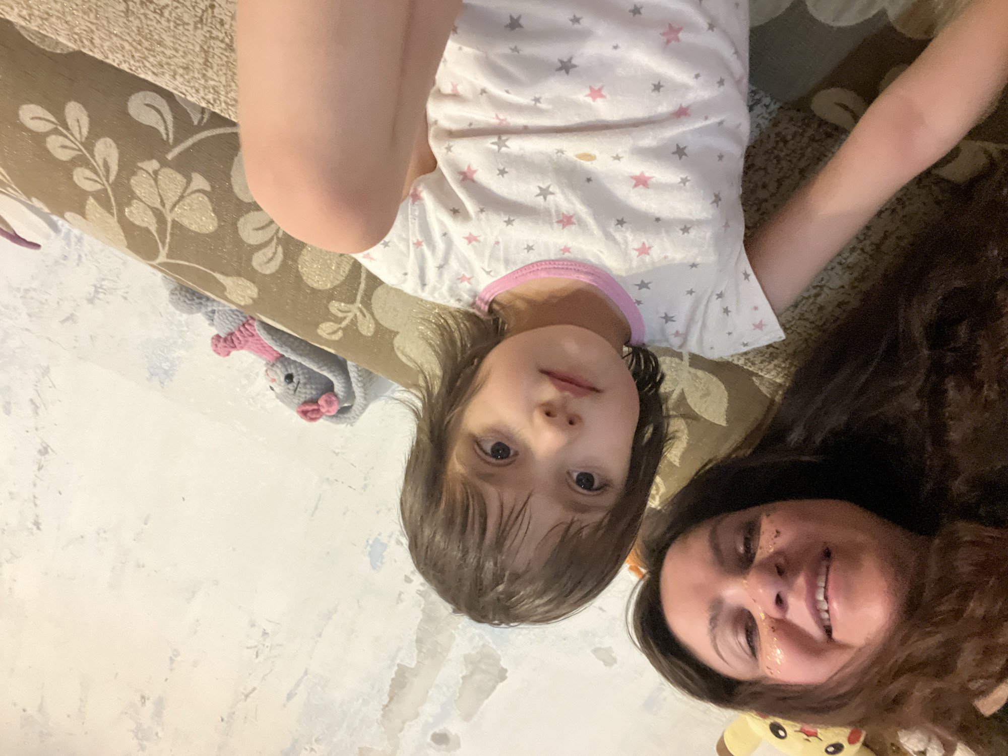 The image shows a woman and a child lying down together indoors. The child is wearing a white shirt with pink and gray stars. The woman has dark hair and is smiling. A gray stuffed rabbit with a pink bow is visible on the left side of the image. A Pikachu stuffed toy is partially visible on the bottom right. The background includes a patterned couch or headboard and a white wall. The location is likely residential. There is no information to confirm this location is in the city of Noviy, Russia.