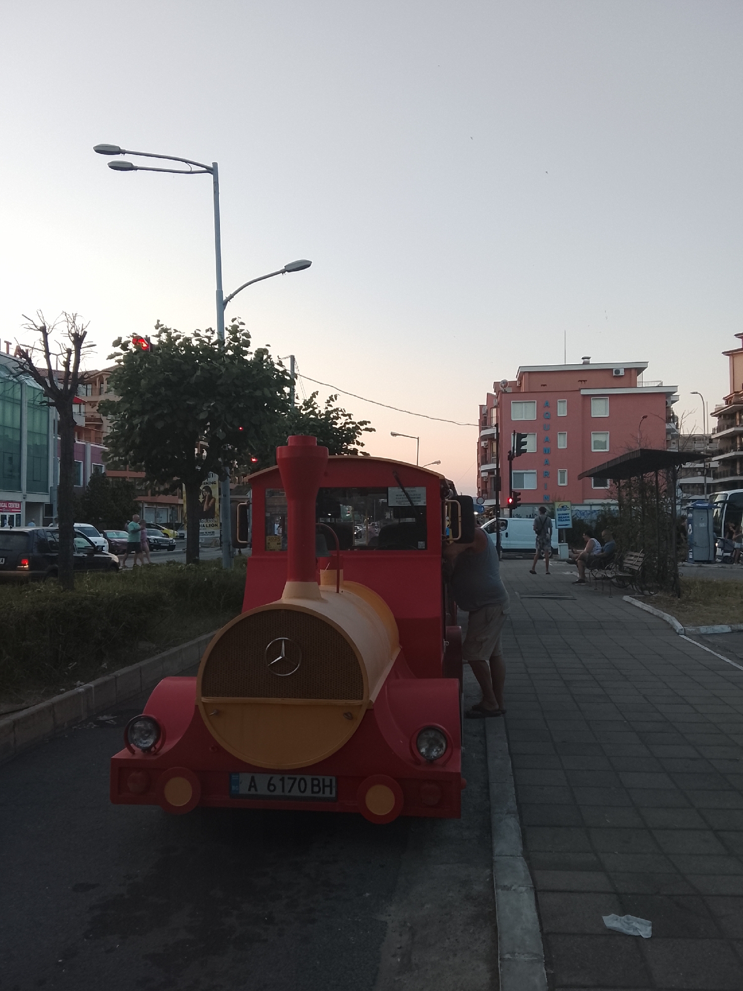 The image depicts a street scene in Nesebar, Bulgaria, near the Aquamarin hotel. In the foreground, a red and yellow toy train, possibly for children's rides, is parked on the road. The train has a Mercedes-Benz logo on its front. A man in a grey tank top and beige shorts stands beside the train, appearing to be interacting with it. A license plate on the train reads "A 6170 BH".
To the right of the train, a paved pedestrian walkway runs alongside the road. Several people are visible on the walkway: a man and a woman walking, and a group of people sitting on a bench under a canopy. Further down the walkway, there is a bus stop with a shelter and a white portable toilet.
The background features buildings, including the pink Aquamarin hotel with "AQUAMARIN" written vertically on its facade. Trees and streetlights are also present, indicating an urban environment. The sky is overcast, suggesting it is either early morning or late evening. The overall impression is of a commercial or tourist area.