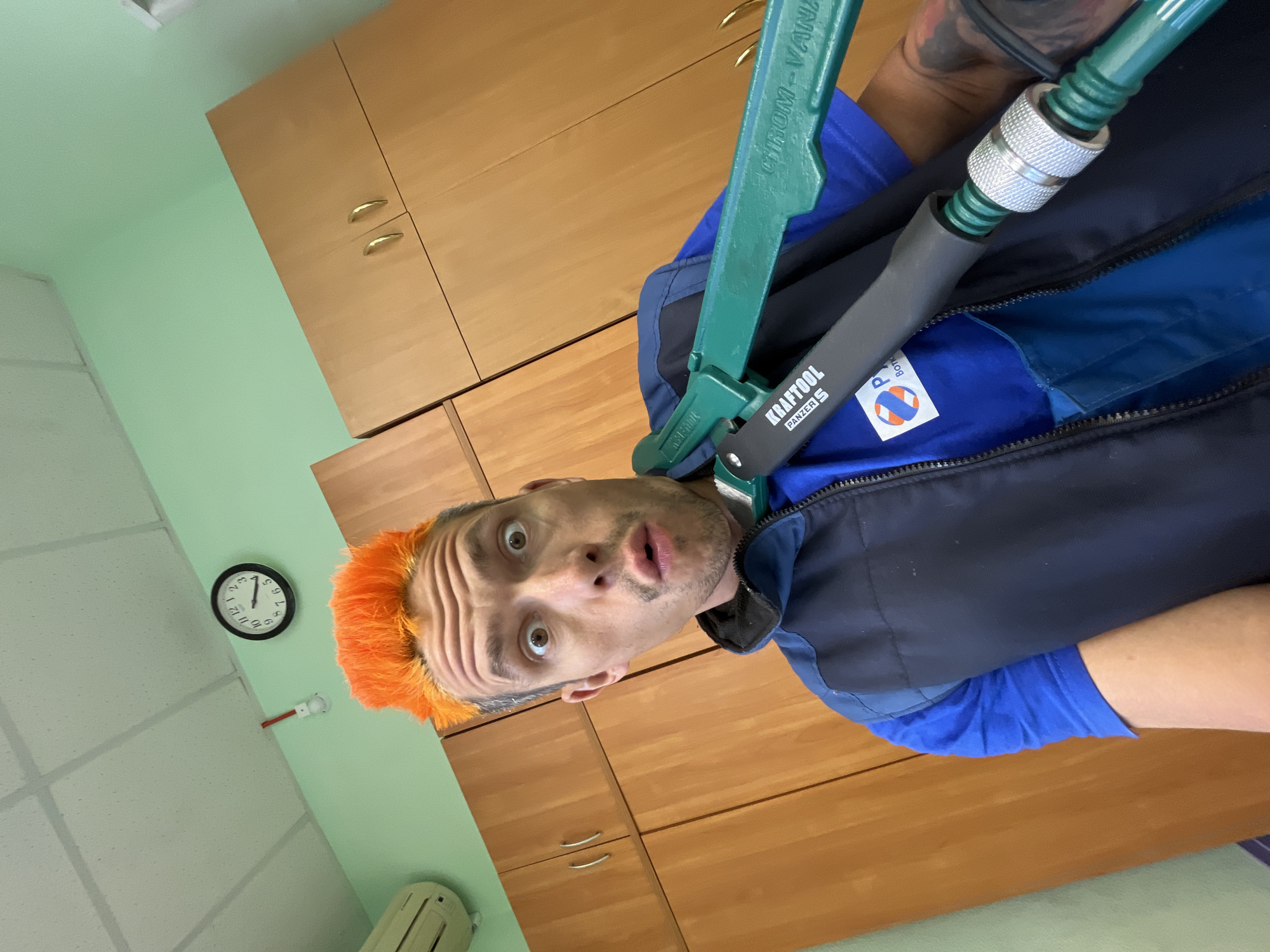 A selfie-style image captures a man indoors in what appears to be an office setting. The man has bright orange hair and is wearing a blue top with a navy vest. He holds a green metal tool, identifiable as a KRAFTOOL PANZER S pipe wrench, against his neck. The background consists of light brown wooden cabinets and a light green wall. A round analog clock is visible on the wall showing approximately 1:05. This image was taken in the city of Chaikovsky, Russia.