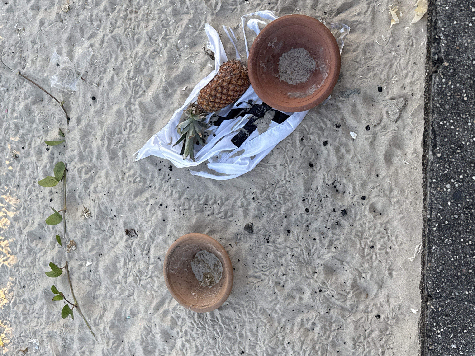 An aerial shot captures a sandy surface with some small footprints. On the left, a terracotta bowl filled with sand and water sits amidst scattered debris. To the right, a pineapple rests on a white plastic bag, accompanied by another terracotta bowl filled with sand. A vine with green leaves snakes across the upper portion of the frame. The bottom of the image shows a rough asphalt surface. The scene appears to be a beach in Niterói, Brazil, possibly Icaraí beach, given the common practice of offering fruits and small terracotta bowls during religious rituals performed near the ocean.