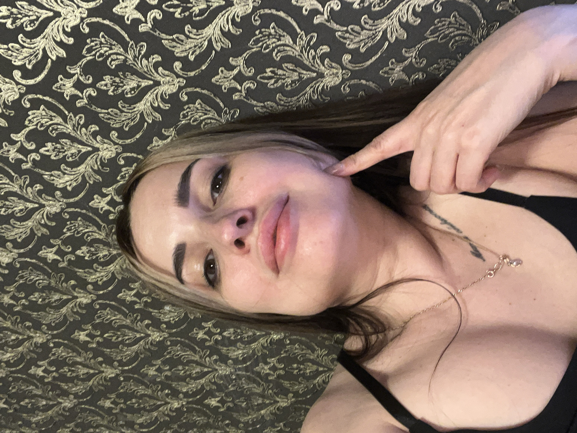 A woman with highlighted brown hair lies supine on a patterned surface, possibly a bedspread or wallpaper, in an indeterminate location. She is wearing a black top. Her right hand is raised, with her index finger pointing towards her right cheek. She has a necklace with a small pendant. The backdrop is dark with gold floral patterns.