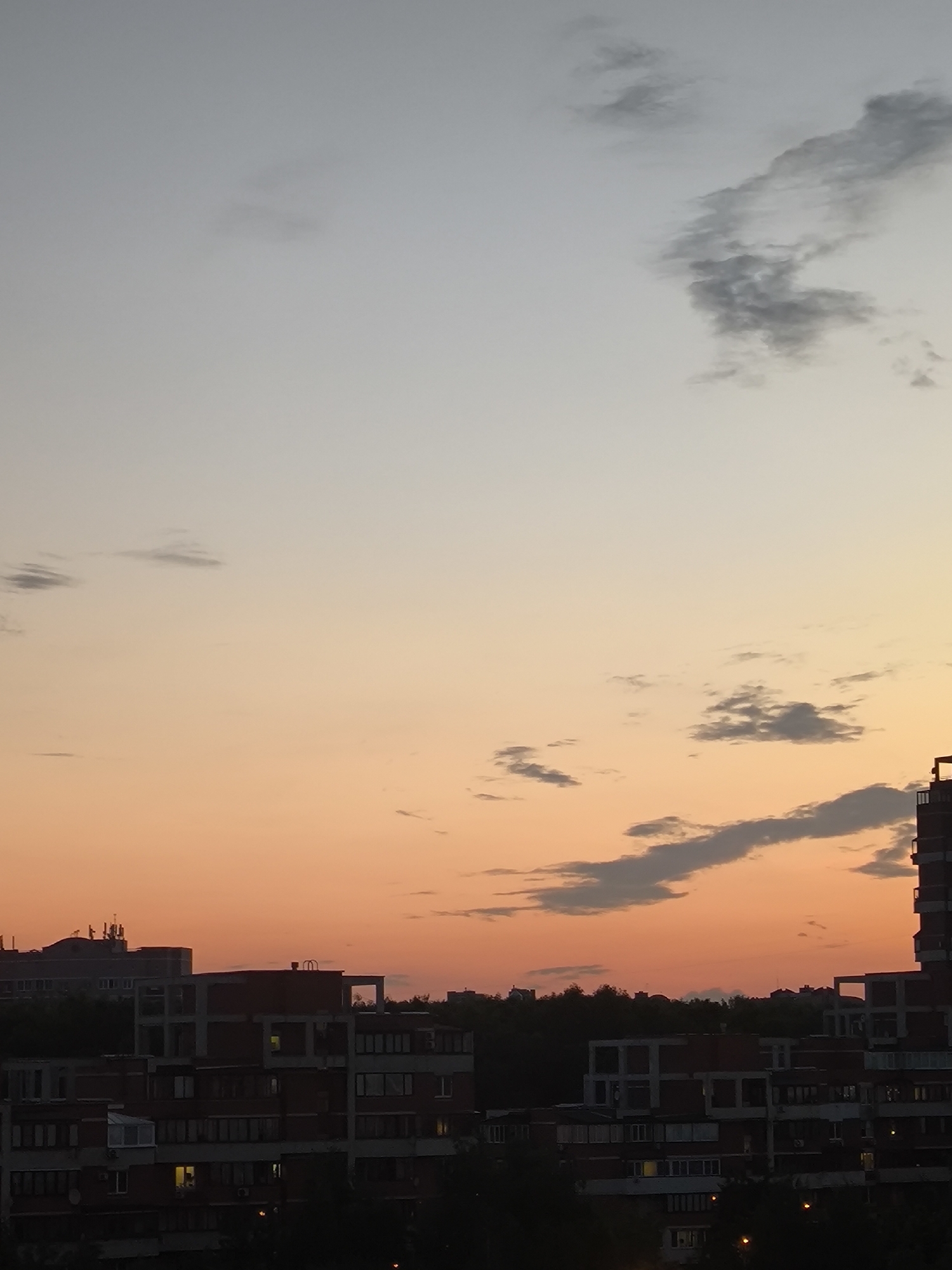 A wide-angle, vertical, outdoor shot captures a twilight sky over an urban landscape. The sky transitions from a pale gray at the top to a warm orange near the horizon. Scattered clouds are visible, some dark gray and others lighter. In the foreground, the silhouettes of several multi-story apartment buildings are prominent against the dusky sky. The buildings appear to be constructed with concrete and brick, with visible windows. Some windows are illuminated, suggesting indoor activity. The dark mass of trees is visible behind and between the buildings, hinting at a dense urban setting. The overall impression is of a residential area at dusk.
