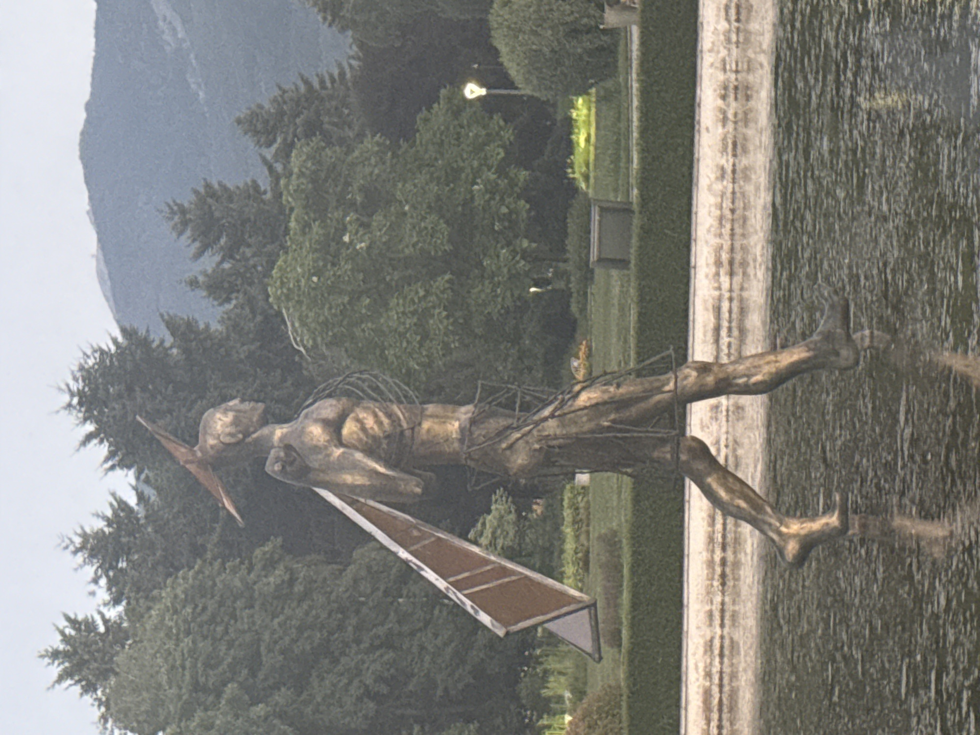 The image features a bronze statue of a man in motion, possibly a runner or a dancer, with a sharp object extending from his head and a rectangular object held in his arms. The statue is positioned in a garden with a fountain or water feature. In the background, there are lush green trees and a large, steep mountain under a cloudy sky. The lighting suggests it is daytime, possibly overcast. The overall scene appears to be a public park or garden in a mountainous region. The statue's metallic finish and dynamic pose are prominent. The water in the fountain reflects the surrounding environment.