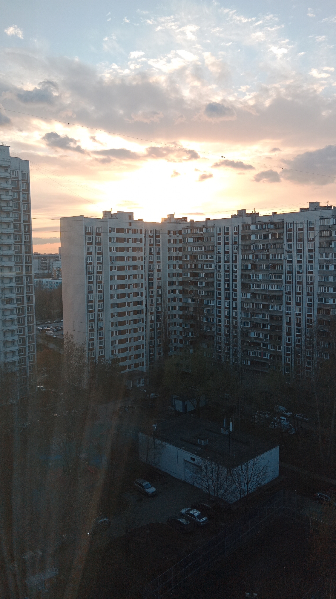 An eye-level, outdoor shot taken in Moscow, Russia, shows apartment buildings under a cloudy, peach-colored sunset.
The buildings, constructed in a gray-white, appear to be Soviet-era apartment blocks with multiple stories and windows. The sky is filled with clouds and a setting sun, creating the peach-colored atmosphere. At ground level, trees, other buildings and parked cars can be seen. The scene is illuminated by the sunset, which also casts shadows.