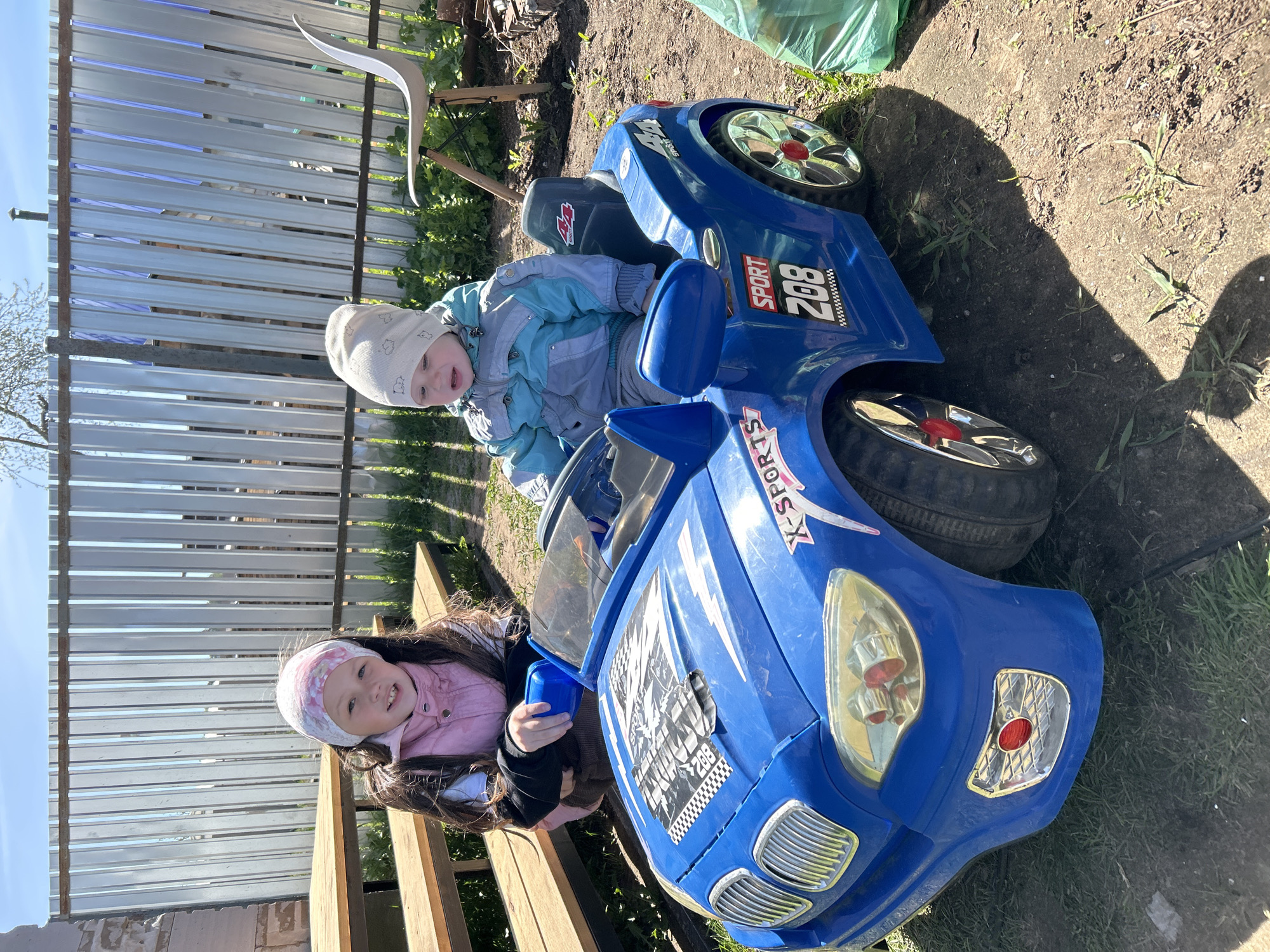 Two children are playing on a blue toy motorcycle in a backyard. A young girl with dark hair and a pink headband sits in the front, while a toddler with a white hat is seated behind her. The motorcycle has "X-Sports" and "208" printed on its sides. The setting is a grassy area with a fence in the background. The location is Chaikovsky, Russia.