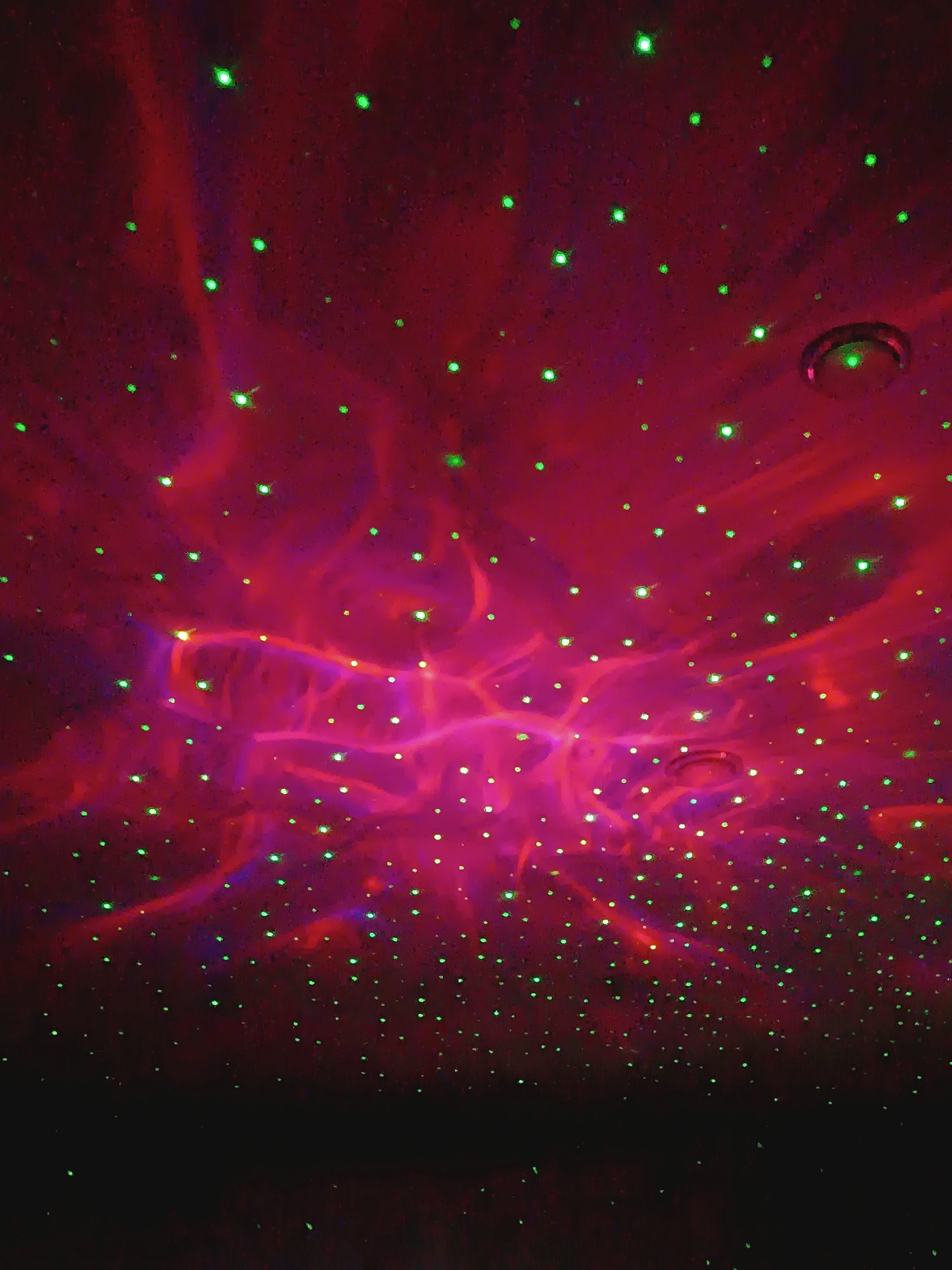 The image shows a dark room lit by a laser projector. Green laser dots are scattered across a red and purple nebula-like pattern projected on the ceiling. The lower portion of the image is dark, suggesting the floor. A circular object, possibly a ceiling fixture, is visible amidst the projected pattern. The absence of people or specific details makes identifying the exact location in Chaikovsky, Russia, impossible.