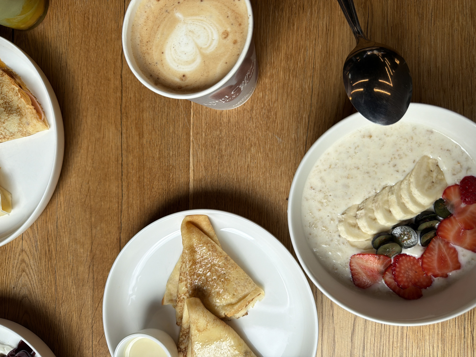 The image depicts a wooden table with several breakfast items. In the foreground, a white bowl contains oatmeal topped with sliced strawberries, blueberries, and bananas. A metal spoon rests on the rim of the bowl, its surface reflecting the overhead lighting. To the upper right of the bowl, a paper cup of coffee with latte art is visible. Above the coffee cup and to the left, a white plate holds folded crepes and a small white dish with a creamy sauce. A sliver of a green beverage container is visible in the top right corner. The overall scene is a top-down view of a breakfast spread on a textured wooden surface, suggesting a cafe or restaurant setting. There is no visual information to suggest this location is in Saint Petersburg, Russia.