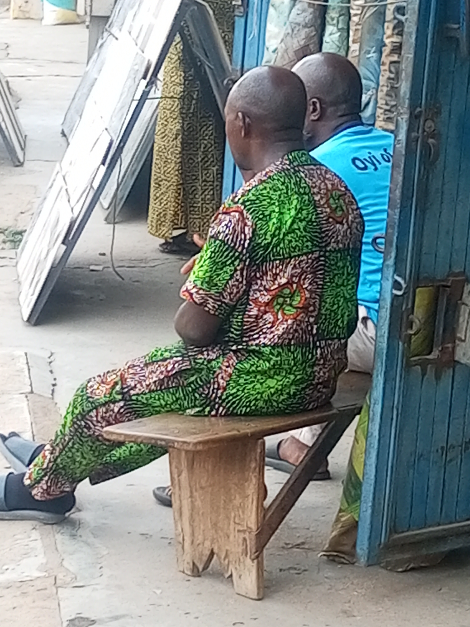 In Maiburiji, Nigeria, two men sit on a wooden bench outside a building with a blue metal door. The man in the foreground wears a matching green and brown patterned shirt and pants. The second man wears a light blue shirt with the words "Oyi of" printed on the back. Several framed items lean against a building to the left. Other individuals are partially visible in the background.